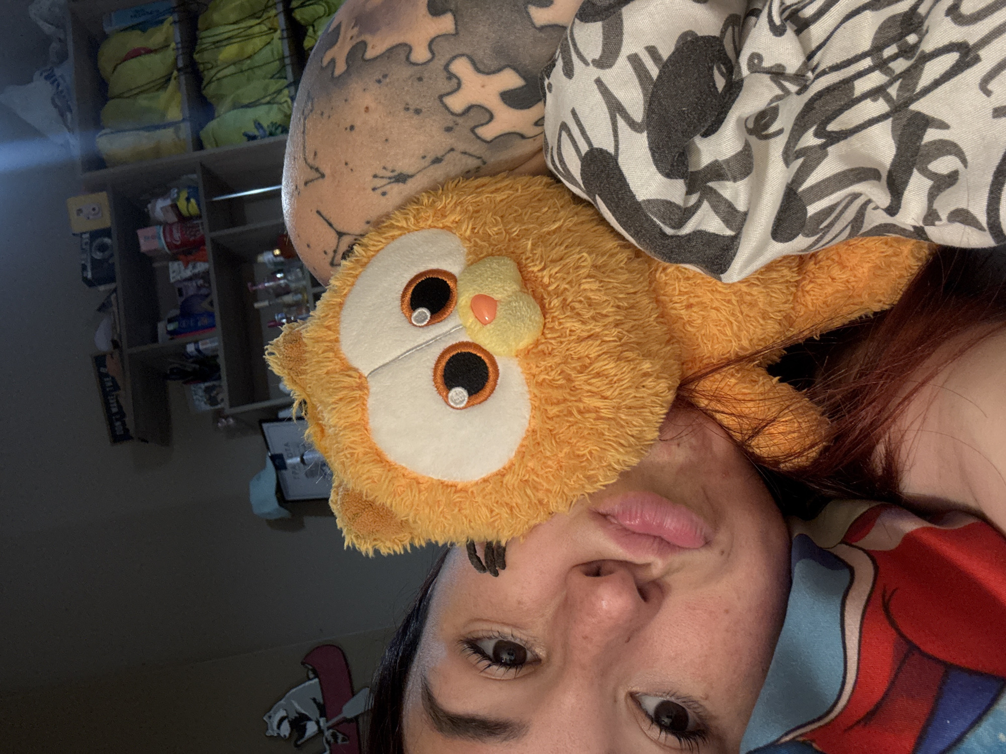 A selfie photograph shows a woman lying down, with an orange owl plush toy positioned on her forehead. Her face is partially obscured by the toy. She has brown eyes and dark hair. Her skin appears to be fair and textured. She is next to a pillow that has a black and white patterned pillowcase. In the background, there's a shelf containing various items. The location in Ruse, Bulgaria, cannot be determined based on the information in the image.