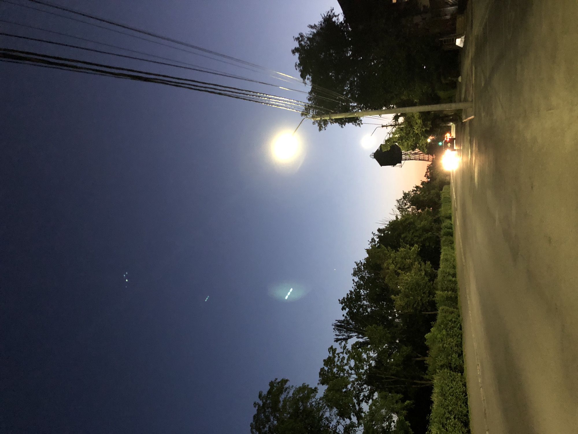 An outdoor, eye-level, medium shot shows a street at dusk in an urban area. The sky is a deep blue, fading to a lighter shade near the horizon. Several light sources are visible in the sky, possibly stars or atmospheric phenomena. Power lines crisscross the upper right portion of the frame. A streetlamp with a bright yellow glow illuminates the scene from the right side. On the left, a row of green bushes lines the street, followed by a line of trees. In the distance, a car with its headlights on is driving down the street, and a tall structure resembling a water tower stands behind the trees. The road surface is asphalt.