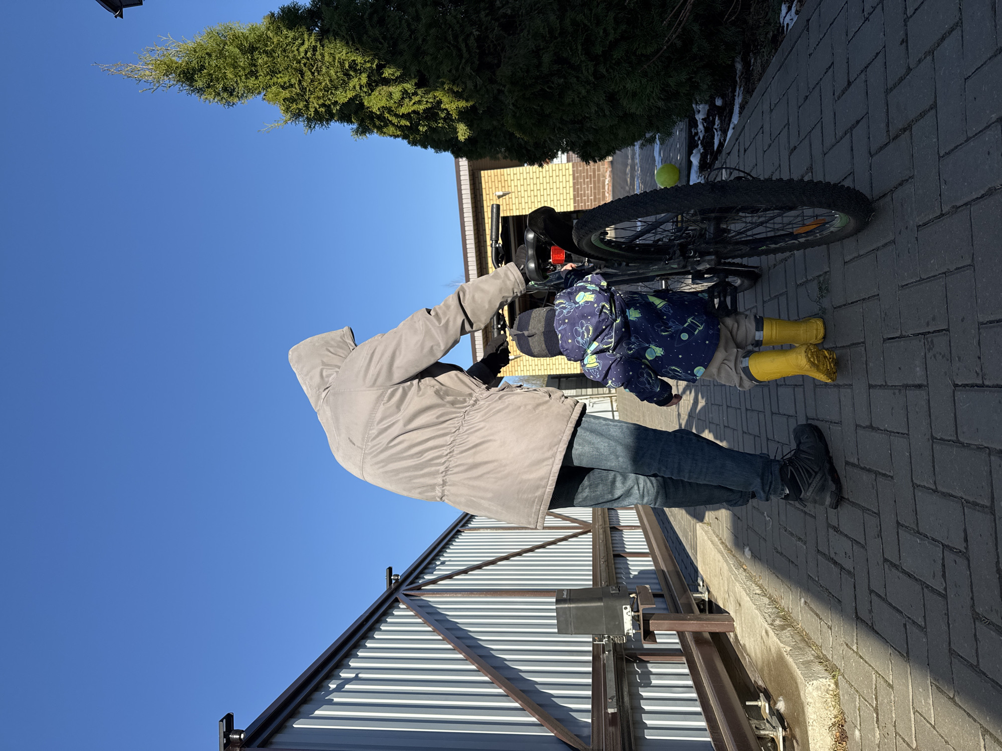 A man in a grey hooded coat and blue jeans stands next to a child in a blue and green patterned jacket and yellow boots. They are both looking at a black bicycle, which is partially visible in the frame. The scene takes place outdoors on a brick pathway. Behind them is a grey metal gate with a mechanical operator. Further back, there is a green bush, a brick building, and a clear blue sky. The location appears to be a residential area. While an Izhevsk location is suggested, there is insufficient detail for a definitive geolocation.
