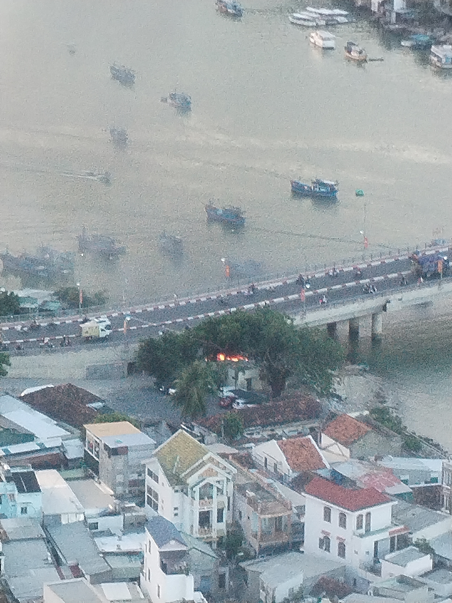 The scene depicts an aerial view of Bac Nha Trang Ward, Vietnam. The water in the foreground hosts various boats, ranging in size. A bridge, marked with alternating red and white sections, spans the water, carrying vehicular traffic and pedestrians. Beneath the bridge, support columns are visible.
Dense clusters of residential buildings with varied roof colors dominate the lower portion of the frame. Building styles vary, with some featuring multiple stories and distinct architectural elements. Mature trees intersperse with the buildings.