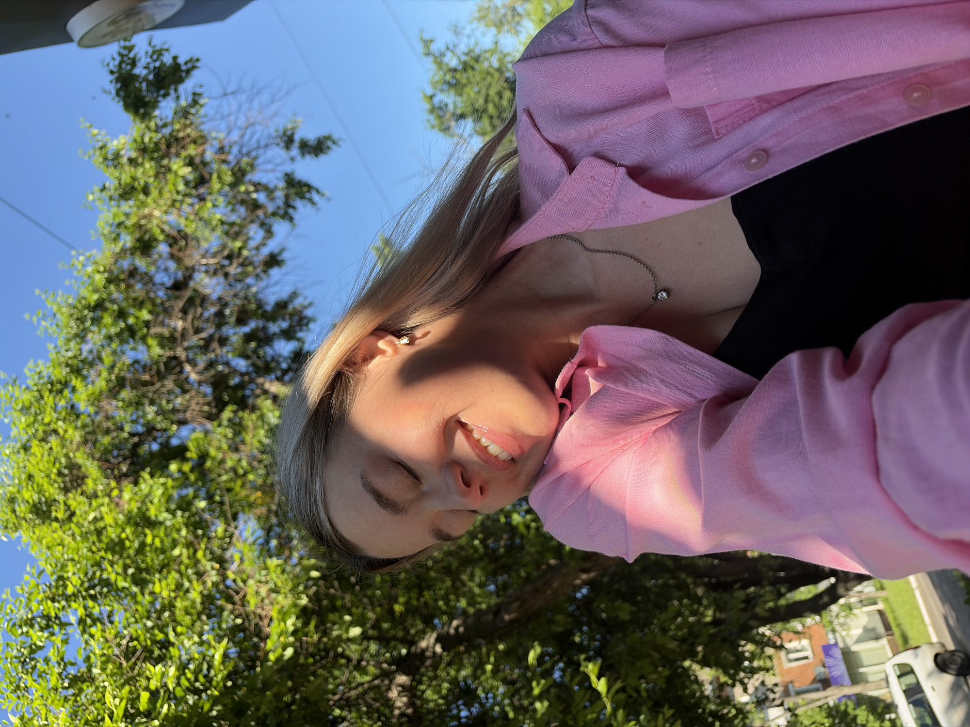A young woman with blonde hair is pictured outdoors, in what appears to be a residential area of Chaikovsky, Russia. She is wearing a pink unbuttoned shirt over a black top, and a silver necklace with a small pendant. Her eyes are closed, and she has a serene expression, suggesting she is enjoying the sunshine which casts a warm glow on her face. The background is dominated by lush green trees and a clear blue sky, with hints of buildings and a white van visible on a street in the lower left corner. The scene appears to be a candid moment captured on a bright, sunny day.