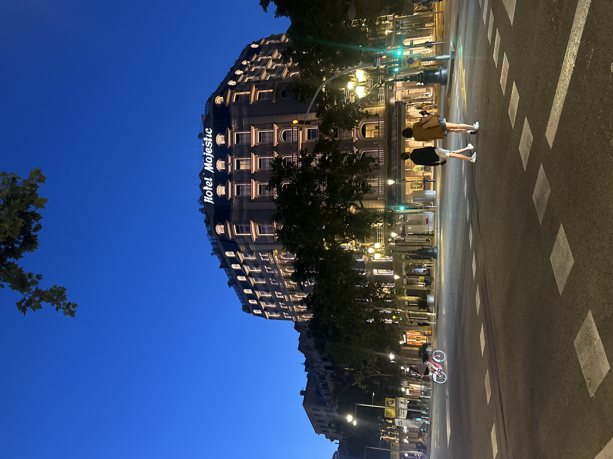 The image shows a street scene at dusk in Barcelona, Spain, in front of the Hotel Majestic. The hotel, a grand, multi-story building with ornate architecture and lit windows, is prominent on the right side of the frame. Its name, "Hotel Majestic," is illuminated in script on the rooftop. Two people, a man and a woman, are walking across a crosswalk in the foreground, with their backs to the camera. They are dressed casually, with the man in a dark shirt and white shorts, and the woman in a brown jacket and denim skirt. A bright pink bicycle with a rider is visible in the background on the left, moving down the street. The street is wide, with streetlights and traffic signals illuminating the scene. Trees line the sidewalk in front of the hotel. The sky is a deep blue, indicating twilight.