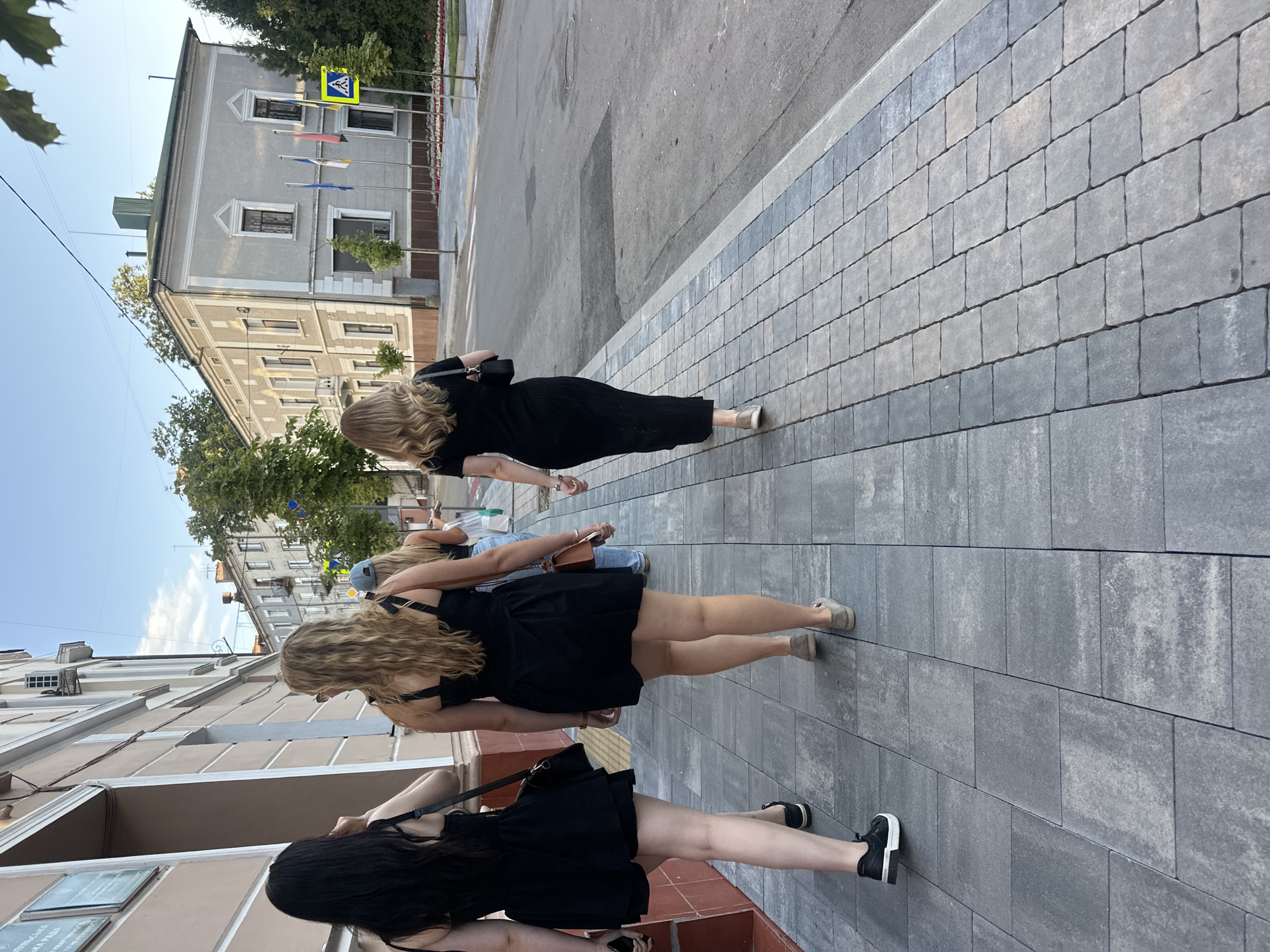 A low-angle medium shot shows four women walking along a sidewalk on a sunny day in Ternopil, Ukraine. The women are dressed in black and walk away from the camera. The woman on the far left has long black hair and wears a black dress and black and white sneakers. The woman next to her has long blonde hair and wears a black dress and tan slip-on shoes. The third woman has blonde hair and wears a blue baseball cap and a blue shirt. The fourth woman is partially obscured and wears a black top and denim shorts. The sidewalk is paved with grey rectangular stones. A building with a light brown facade is on the left, and a grey building with a green roof is on the right. A street with a crosswalk sign is visible to the right of the buildings. The sky is clear and blue.