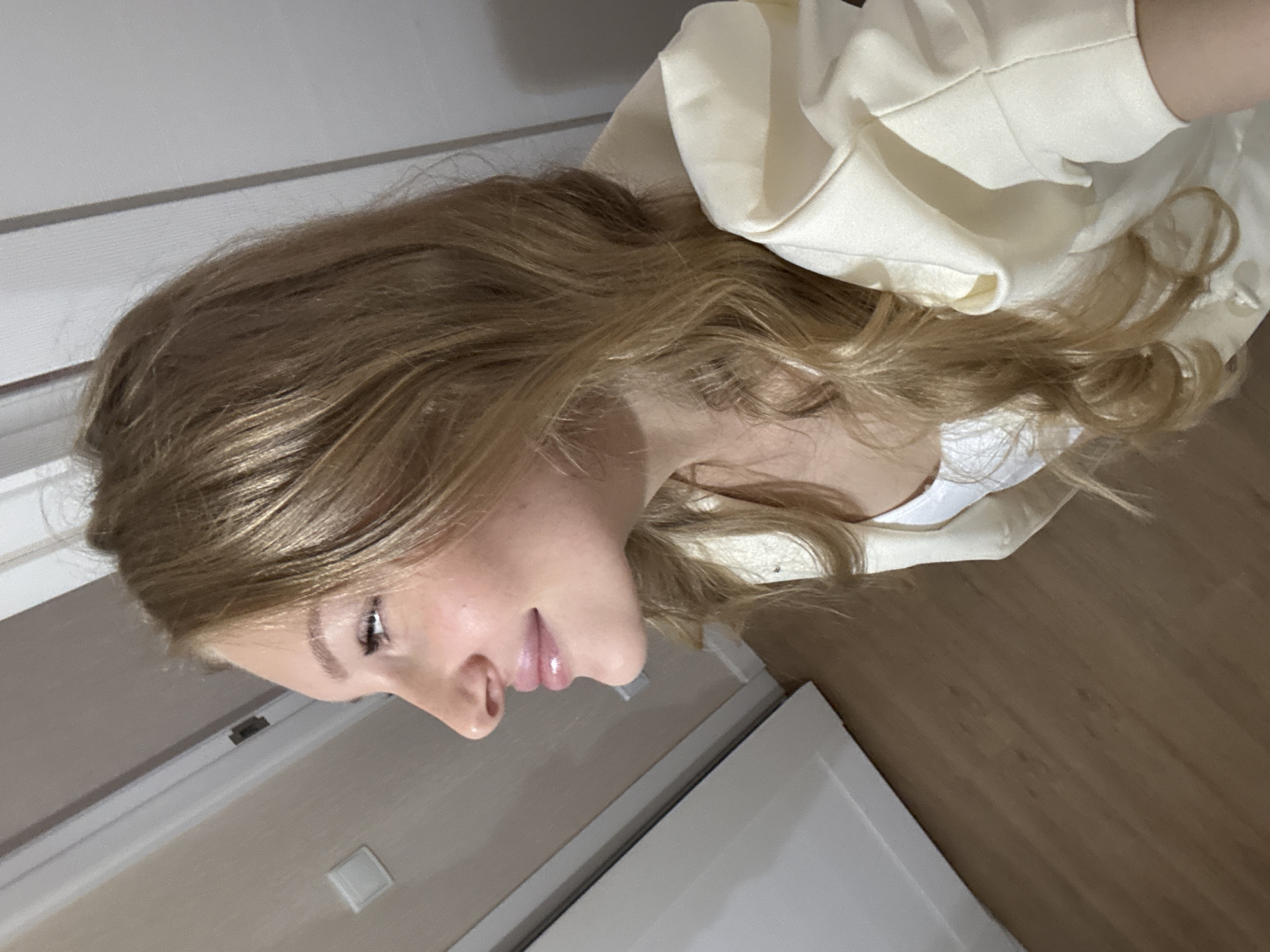 A young woman with blonde hair and fair skin is shown in profile, looking to the left. She is wearing a white tank top and a light yellow jacket with puffed sleeves. Her lips are slightly parted and appear to be glossy. The background consists of a wooden floor, a light-colored wall with a white door or cabinet, and a white wall. The lighting suggests an indoor setting, possibly a home or apartment in Perm, Russia, given the context.