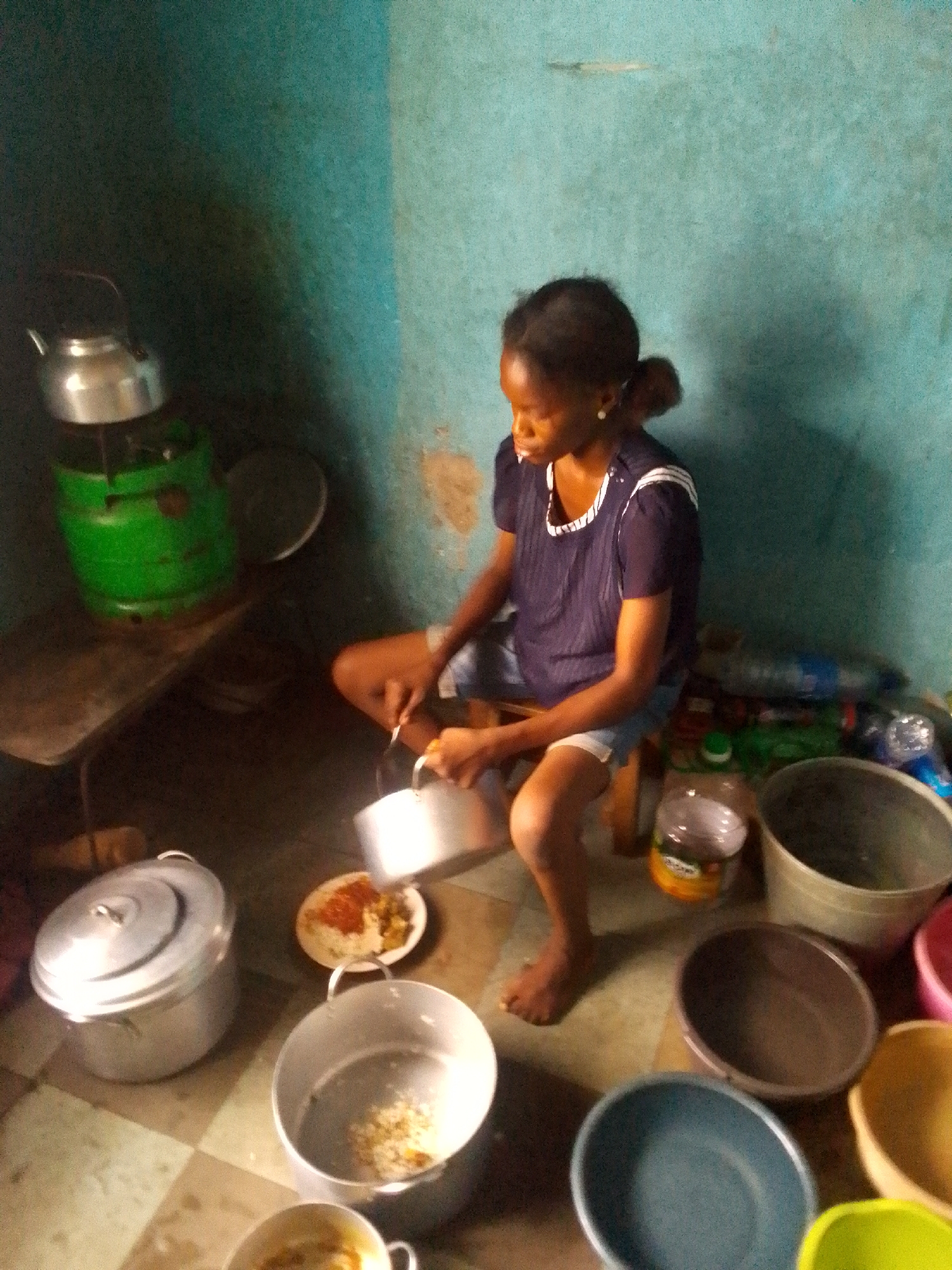 A woman is squatting in a corner of a room, pouring food from a pot into a plate that is already filled with food. There is a larger pot on the floor next to her and several empty bowls. Behind her are some bottles and a shelf with a metal kettle on top. The room has blue walls and a tiled floor. The location is unknown.