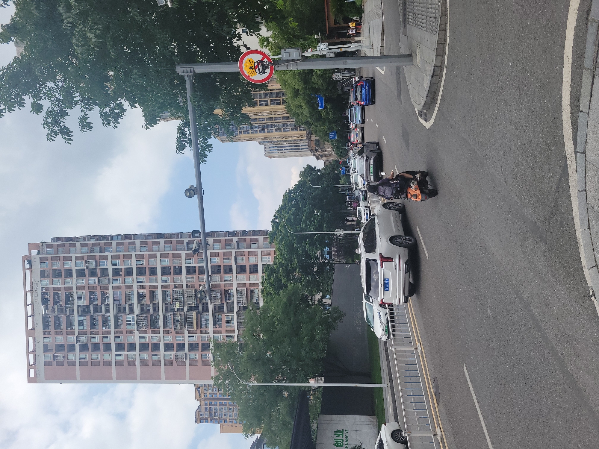 The image depicts a street scene in Yuzhong District, China, under a cloudy sky. A tall, multi-story residential building with numerous balconies and windows dominates the left side of the frame, with shorter, similar buildings visible in the background. A paved road occupies the foreground and middle ground, with traffic moving in both directions. Several vehicles are visible, including white SUVs, sedans, and a blue sedan. A person is riding an orange electric scooter in the right lane of the road, facing away from the camera. A traffic light is visible overhead, as is a regulatory sign with a red circle and a diagonal line over an image of a truck with an explosion, indicating no trucks. Trees line the sides of the road, and a sidewalk with pedestrians is present on the right. A white metal fence separates the road from a grassy area on the left, where a building with Chinese text is partially visible.