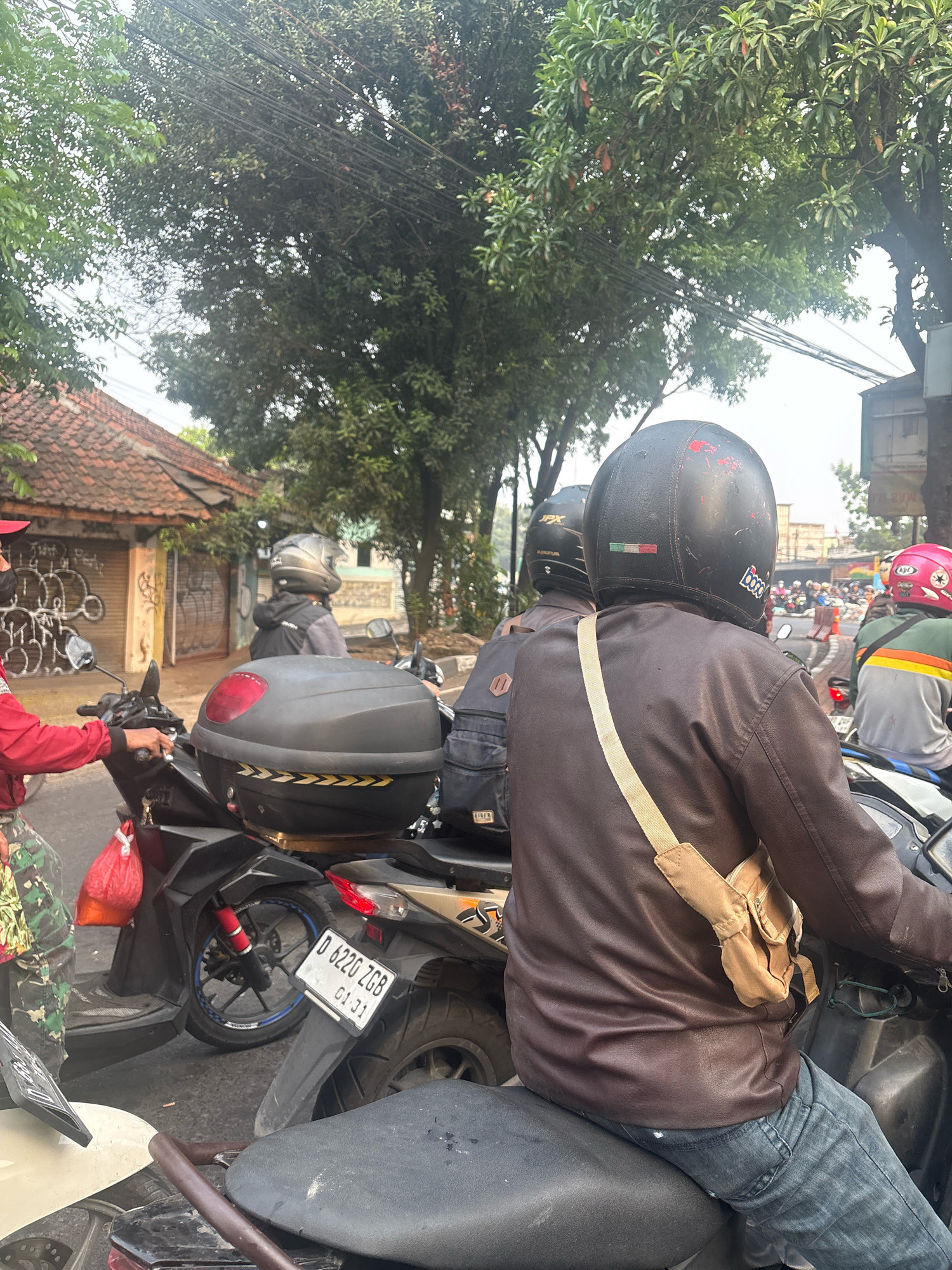 In a street scene in Bandung City, Indonesia, several motorcyclists are stopped in what appears to be traffic. The foreground features a person wearing a brown leather jacket and blue jeans, with a beige crossbody bag. They are seated on a motorcycle with a black storage box. To their left, another person in a red jacket and camouflage pants is visible, holding a red plastic bag. Behind them, several other motorcyclists are present, some wearing helmets. The background includes trees, buildings with tiled roofs, and street infrastructure. The license plate of the motorcycle in the foreground reads "D 6220 ZGB 01-31". The overall atmosphere suggests a typical urban environment with moderate traffic.