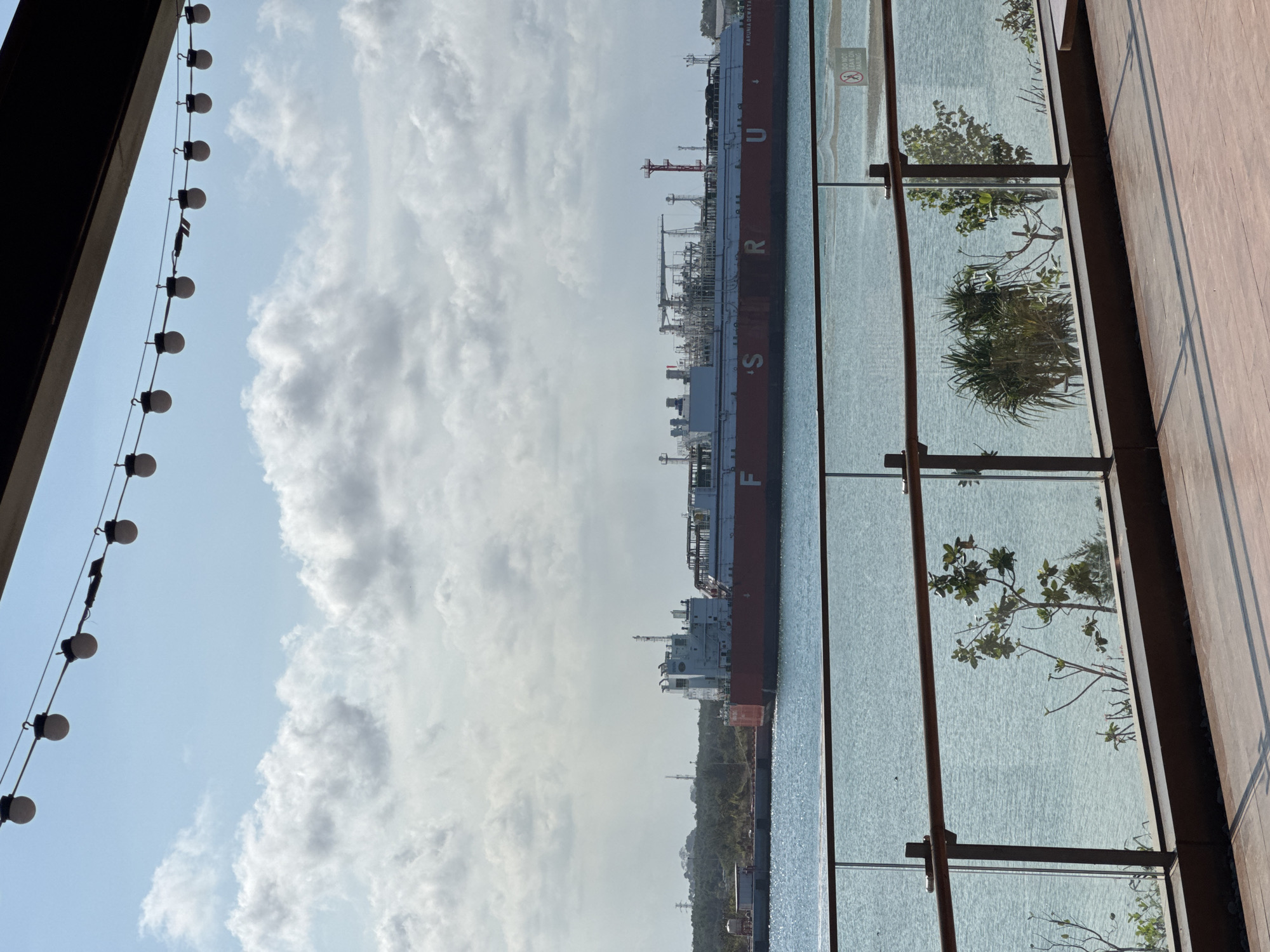 A large, red and white ship, identified by the lettering "KARUNIA DEWATA" and "FSRU" on its side, is docked in the harbor of an unknown city in Indonesia. The ship has an industrial processing plant on its deck. In the foreground, there is a wooden deck with a glass railing. Beyond the railing is the turquoise water of the harbor, with several trees and shrubs visible on the shoreline in the distance. The sky is partly cloudy, with bright sunlight illuminating the scene. The overall impression is one of a busy industrial port with a scenic waterfront.