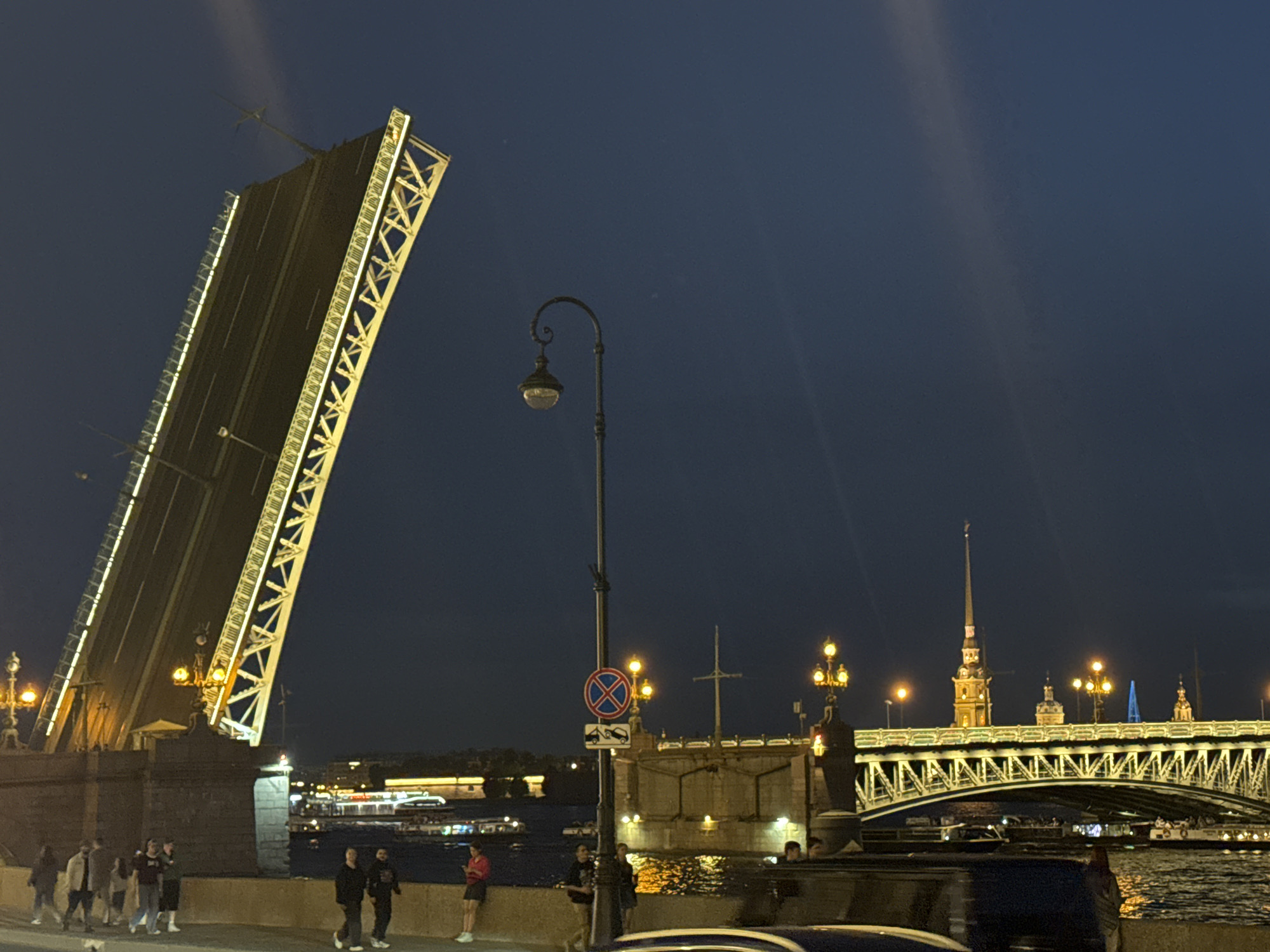 The image captures a nighttime urban scene in Saint Petersburg, Russia, featuring the iconic Palace Bridge (Dvortsovy Most) and Trinity Bridge (Troitsky Most) spanning the Neva River. In the left foreground and midground, the Palace Bridge is prominently displayed with its two central drawbridge spans fully raised, forming a steep 'V' shape against the dark blue sky. These raised spans are brightly illuminated along their edges and structural framework with warm yellow and white lights. Below the raised bridge sections, a stone embankment runs parallel to the river, where approximately 10-15 individuals are observed. They are dressed in casual clothing, some walking in groups, others standing and observing the illuminated bridges and river.
In the center of the image, a tall, ornate black streetlamp with a single light fixture is positioned, and a red circular "No Stopping/Parking" road sign with a tow-away symbol is attached to its pole. To the right of the streetlamp, the Trinity Bridge is visible in the midground, its arched metal truss structure and roadway illuminated with warm yellow lights. Behind the Trinity Bridge, the distinctive spire of the Peter and Paul Cathedral rises into the night sky. The dark waters of the Neva River reflect the city and bridge lights. Several boats, some with their own lights, are visible on the river. The sky is a deep dark blue, showing subtle light streaks from the intense bridge illumination. Parts of vehicles are present in the immediate bottom foreground, suggesting a street alongside the embankment.