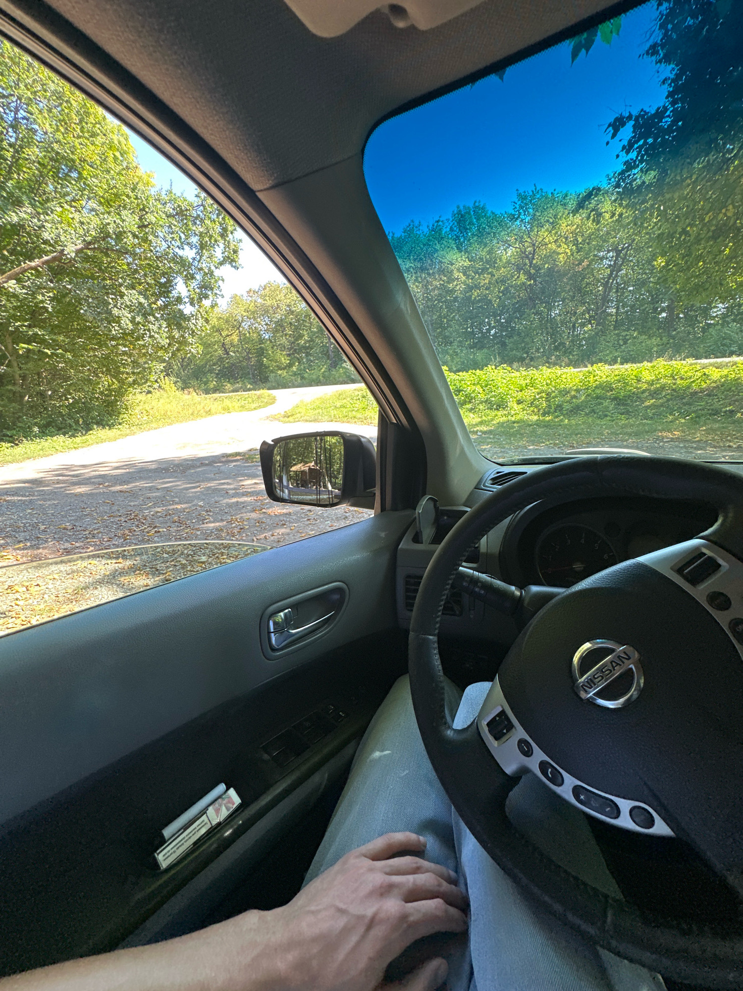 The image is a first-person perspective from inside a Nissan vehicle, looking out the driver's side window and through the windshield. The vehicle is on a gravel road surrounded by lush green trees and foliage, suggesting a rural or natural setting. The sky is a clear, bright blue. In the driver's side mirror, a small, brown, wooden structure, possibly a birdhouse or decorative item, is visible. On the interior door panel, a pack of cigarettes and a single cigarette are placed in a compartment. The steering wheel of the Nissan is in the foreground, with the driver's hand resting on their lap, wearing light-colored jeans. The overall scene depicts a person driving through a scenic, wooded area, likely in or near Shatava, Ukraine, based on the provided location information.