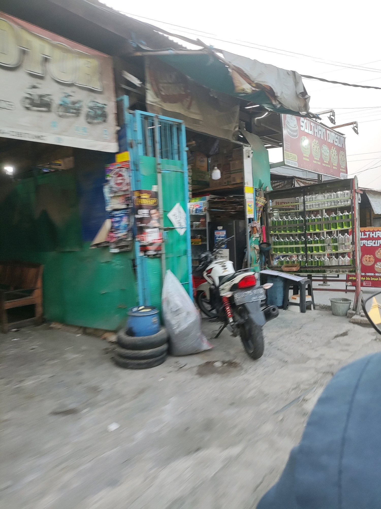 The image shows a street-level view of a shop in Wanajaya, Indonesia. The shop appears to be selling various goods, including what looks like fish in bottles displayed on shelves. There is a motorcycle parked in front of the shop, and several tires stacked near the entrance. A blue barrel is also visible, along with a large grey bag. The shop itself has a green facade and a corrugated metal roof with some tarps. Signs are visible, one with text that appears to be in Indonesian, and another with an image of food. The ground is paved, and the overall environment suggests a commercial area in an Indonesian city. The lighting is overcast, indicating it might be daytime.