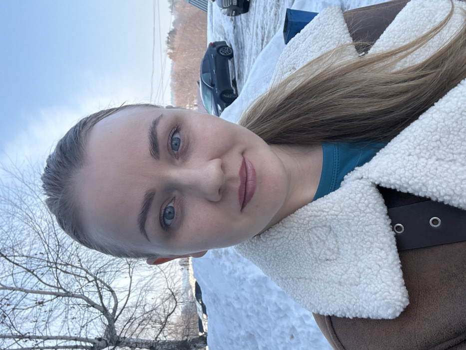 A fair-skinned female subject is depicted in a close-up outdoor portrait, likely a selfie, set against a snowy winter backdrop in Chaikovsky, Russia. The subject, appearing to be in her 20s or 30s, has medium blonde hair pulled back, with a section draped over her right shoulder. She possesses blue eyes, light eyebrows, and is presented with a slight smile, featuring light pink lips. Her attire includes a brown, presumably faux-suede, jacket with a prominent cream-colored sherpa or fleece lining visible on the collar and lapels. Underneath, a teal crew-neck top is visible.
The environment indicates a clear, bright winter day with a blue sky overhead and significant snow accumulation on the ground. To the left of the subject, a bare, leafless deciduous tree is visible. To the right, a paved area, likely a parking lot or roadside, hosts a dark-colored sedan (resembling a Kia Optima) parked diagonally, partially obscured by snow. Another lighter-colored vehicle is partially visible further to the right. A blue rectangular bin or container is situated on the far right. In the distant background, hills or a treeline are discernible, suggesting an open landscape.