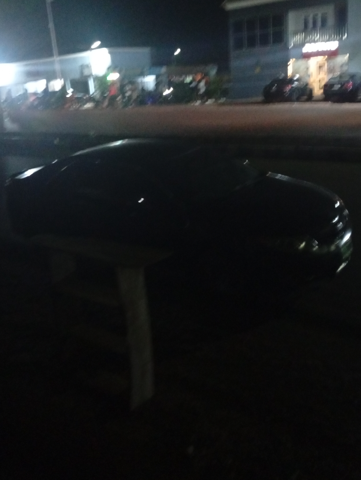 The image depicts a dark outdoor scene, likely at night, in Adankolo, Nigeria. A dark-colored car is positioned in the foreground, partially obscured by the darkness. It appears to be parked on or near a table or stand. In the background, there is a building with lights on, possibly a store or restaurant, with other cars parked nearby. People can be seen near the building, but they are not clearly discernible due to the low light conditions.