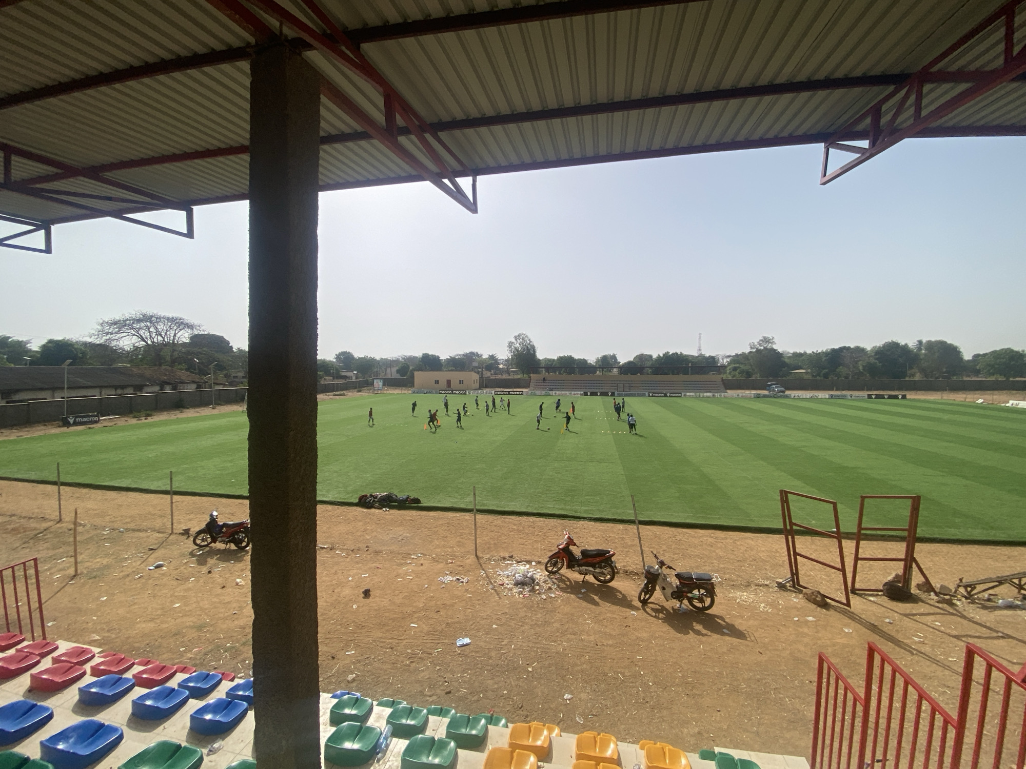The image shows a view of a soccer field and its surrounding area from a partially covered stadium. The field is green with striped mowing patterns, and a group of people are practicing on it. The stadium seating area is in the foreground and is made up of seats in red, blue, green, and yellow colors. The ground surrounding the field and the seating area is mostly dirt. Several motorbikes are parked on the dirt area between the stands and the field. Overhead, a corrugated metal roof structure partially covers the stadium area. The image was taken in Tudun Wada, Nigeria.