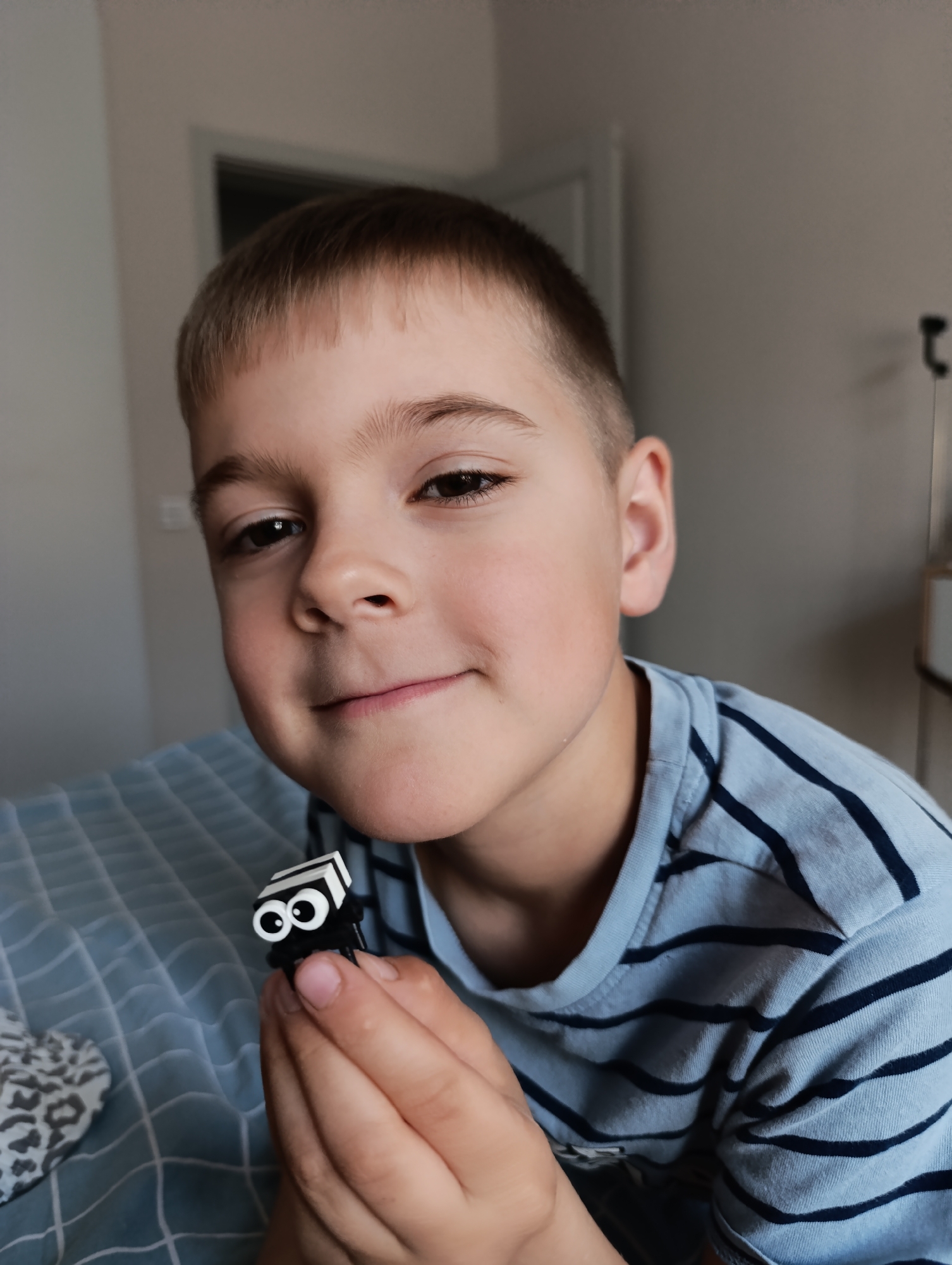 A young boy with short brown hair is holding a black and white toy with googly eyes in his hands. He is smiling slightly. He is wearing a blue and black striped shirt. The background includes a bed with a patterned comforter, a door, and a wall. This image was taken in a residential area, likely indoors, possibly in Chaikovsky, Russia.