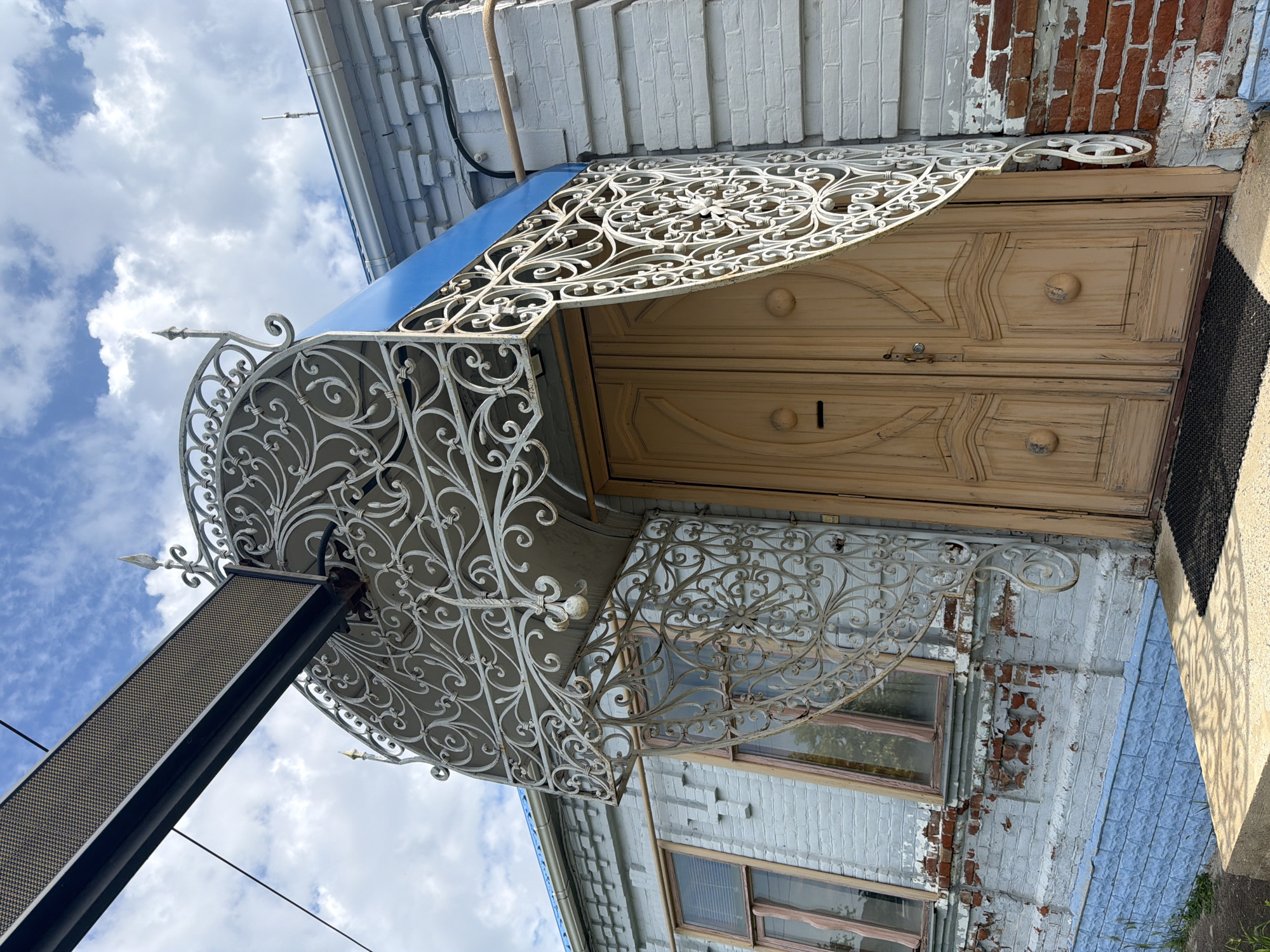 The image shows the entrance to a brick building in Leningradskaya, Russia. The building has a light blue facade with exposed red brick in some areas. The entrance features a double wooden door painted a light brown color. Above the door is an ornate white metal awning with intricate scrollwork. A blue fabric canopy is visible beneath the metal awning. Two windows with wooden frames are on the left side of the entrance. The sky above is partly cloudy.