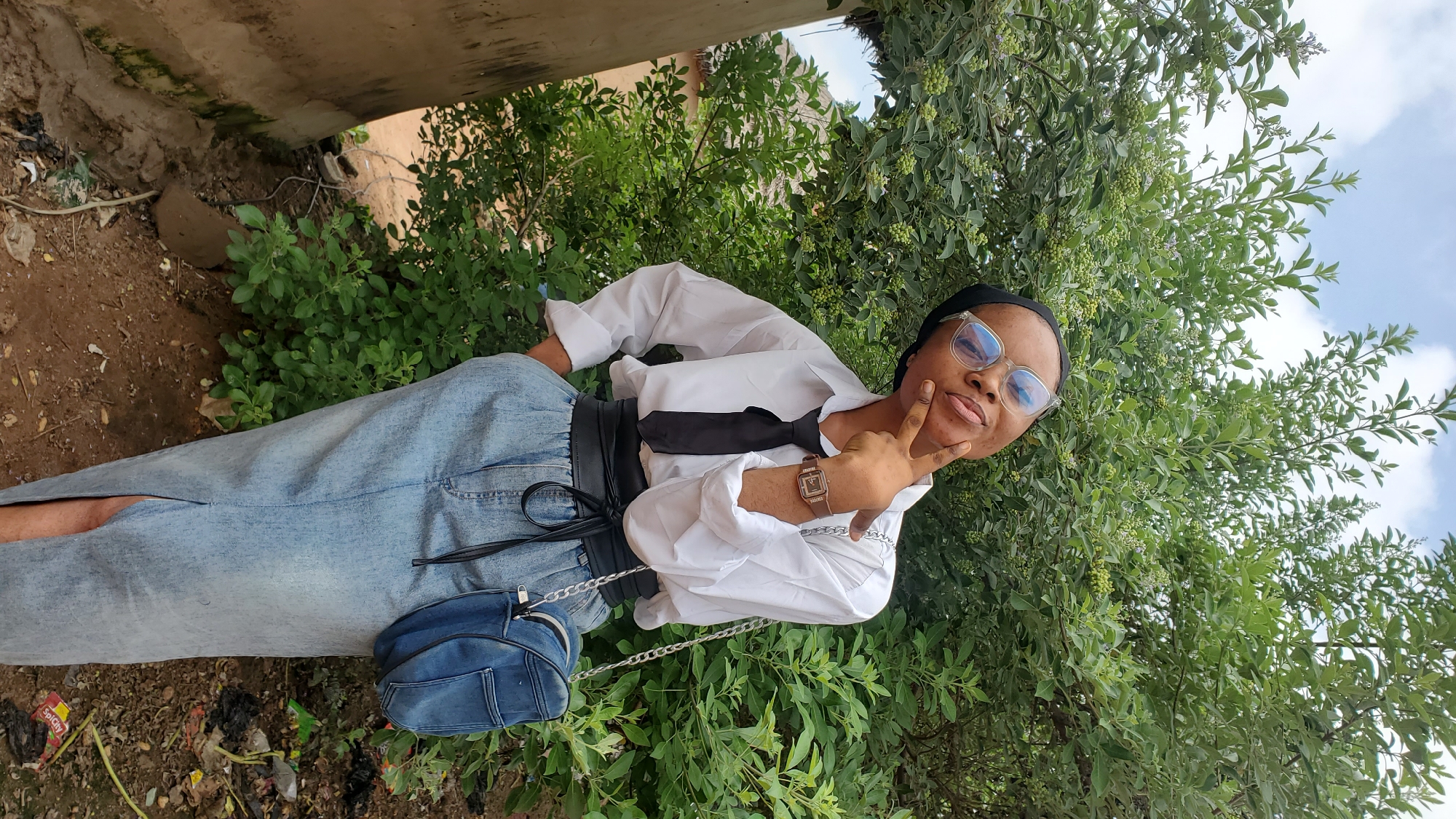 A woman stands outdoors in front of a dense green bush, her body angled slightly towards the viewer. She is wearing a white button-down shirt with a black tie, a black belt cinched at her waist, and a light blue denim skirt with a slit up the front. She has on large, light-rimmed glasses and a black headscarf. Her left hand is on her hip, and her right hand is raised, with her index finger touching her chin, in a thoughtful pose. A small, blue denim purse with a silver chain strap hangs on her right side. The background consists of the leafy green bush and a cloudy sky. To the left of the woman, a portion of a light-colored, textured pillar is visible, with some greenery and dry soil at its base. The ground is covered in dry soil, with scattered leaves and small debris, including a red and yellow wrapper that appears to be from a snack. The scene suggests an outdoor setting, possibly a residential area or a park, within the city of Jalingo, Nigeria.