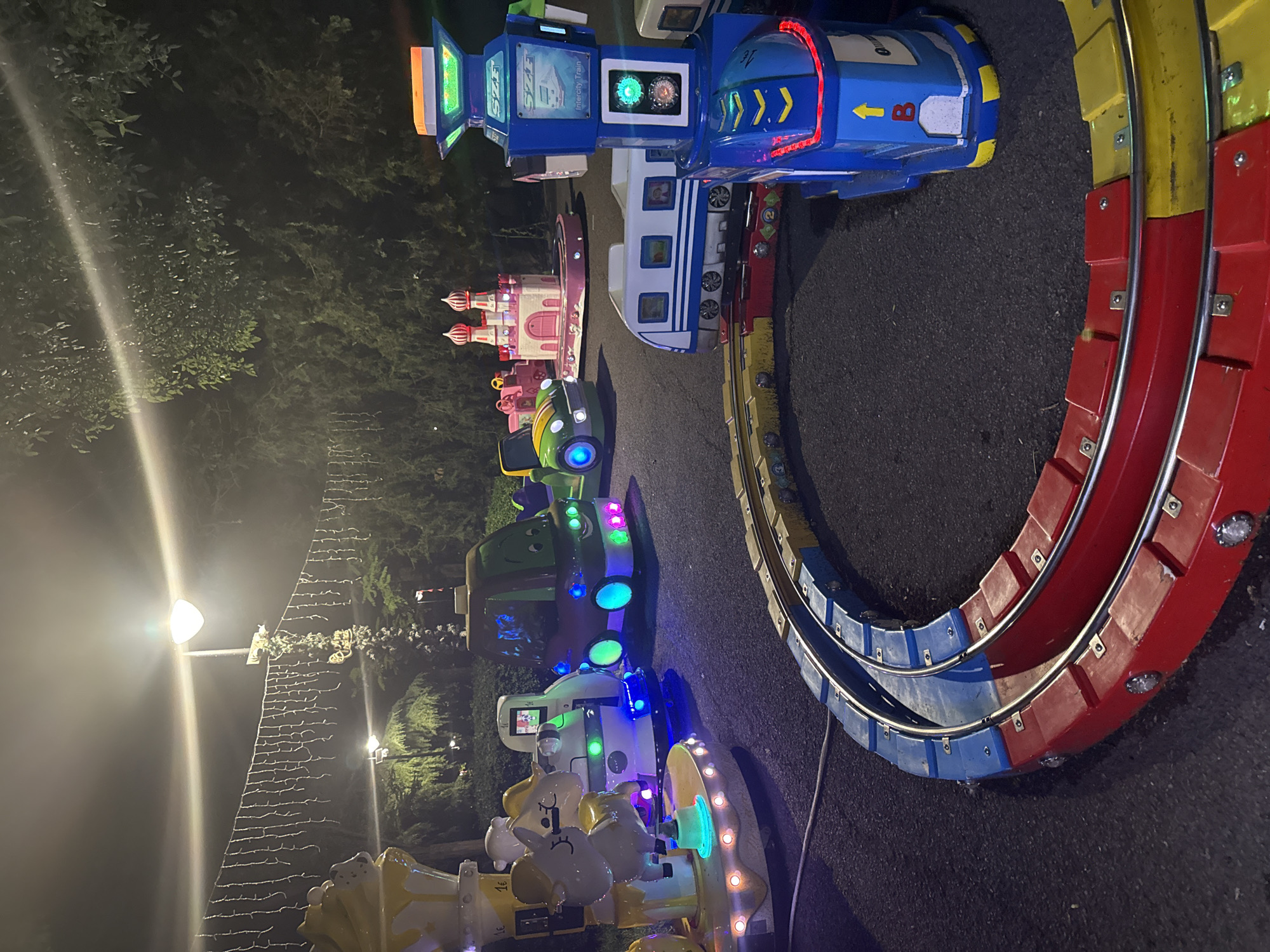 This nighttime image depicts an outdoor children's amusement area, likely in Burgas, Bulgaria, characterized by a line of brightly illuminated coin-operated rides. The setting is dark, with artificial lighting providing the main source of illumination.
The most prominent feature is a blue and white "Intercity Train" ride positioned on a colorful track. The train itself is adorned with numerous lights and features screens, one displaying what appears to be traffic signals with green and red lights. Text visible on the train includes "Intercity Train" and partially, "STAR," along with a directional arrow and the letter "B." The track beneath it is a striking combination of red, yellow, and blue sections, curving sharply to the right, and also features integrated lights.
Behind the train ride, a series of other children's rides are arranged. These include a pink castle-themed ride, a green vehicle, a white vehicle, and a yellow ride featuring cartoonish character faces, all equipped with various colorful LED lights that glow vibrantly in the darkness.
No people are visible in the image, suggesting the rides are either temporarily inactive or the area is quiet. In the background on the left, a strong, almost starburst-like light emanates from a street lamp, casting bright streaks that might indicate mist or haze. Dark foliage from trees and bushes frames the upper and left parts of the image, contributing to the nighttime atmosphere. Some decorative string lights are also visible in the mid-ground on the left. The ground is dark, appearing to be asphalt or pavement. The scene evokes a playful and vibrant mood typical of a nighttime entertainment venue.