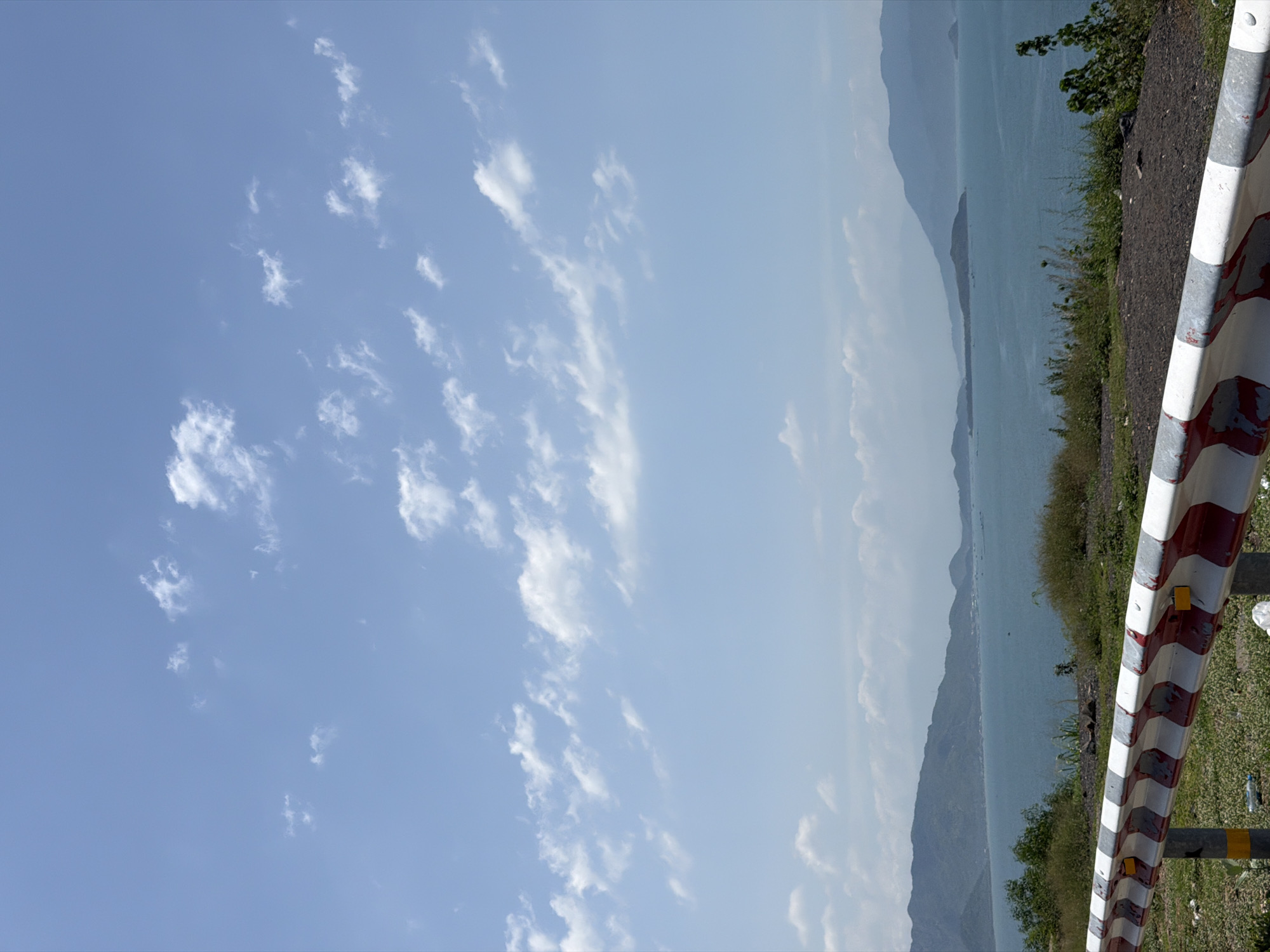 This image presents a panoramic coastal landscape viewed from an elevated perspective in Bac Nha Trang Ward, Vietnam. The foreground features a white and red chevron-patterned road barrier, running horizontally across the lower section, partially obscured by sparse dry grass, dark soil, and visible litter (plastic bottles). Beyond the barrier, a vast expanse of calm, deep blue-green water, likely a bay, dominates the midground. Scattered across the water are several small, dark specks, indicative of distant boats or marine structures. Along the midground's distant shores and within the bay, multiple vegetated landmasses, appearing as islands or peninsulas, rise from the water. Closer to the left-center, a faint line of development, possibly a coastal town or village, is visible against the foothills. The background is characterized by a series of hazy, vegetated mountains extending across the horizon. The upper two-thirds of the image consist of a clear, light blue sky with widely scattered white cumulus clouds. The scene is brightly illuminated, suggesting midday lighting conditions, and no human figures or animals are present.