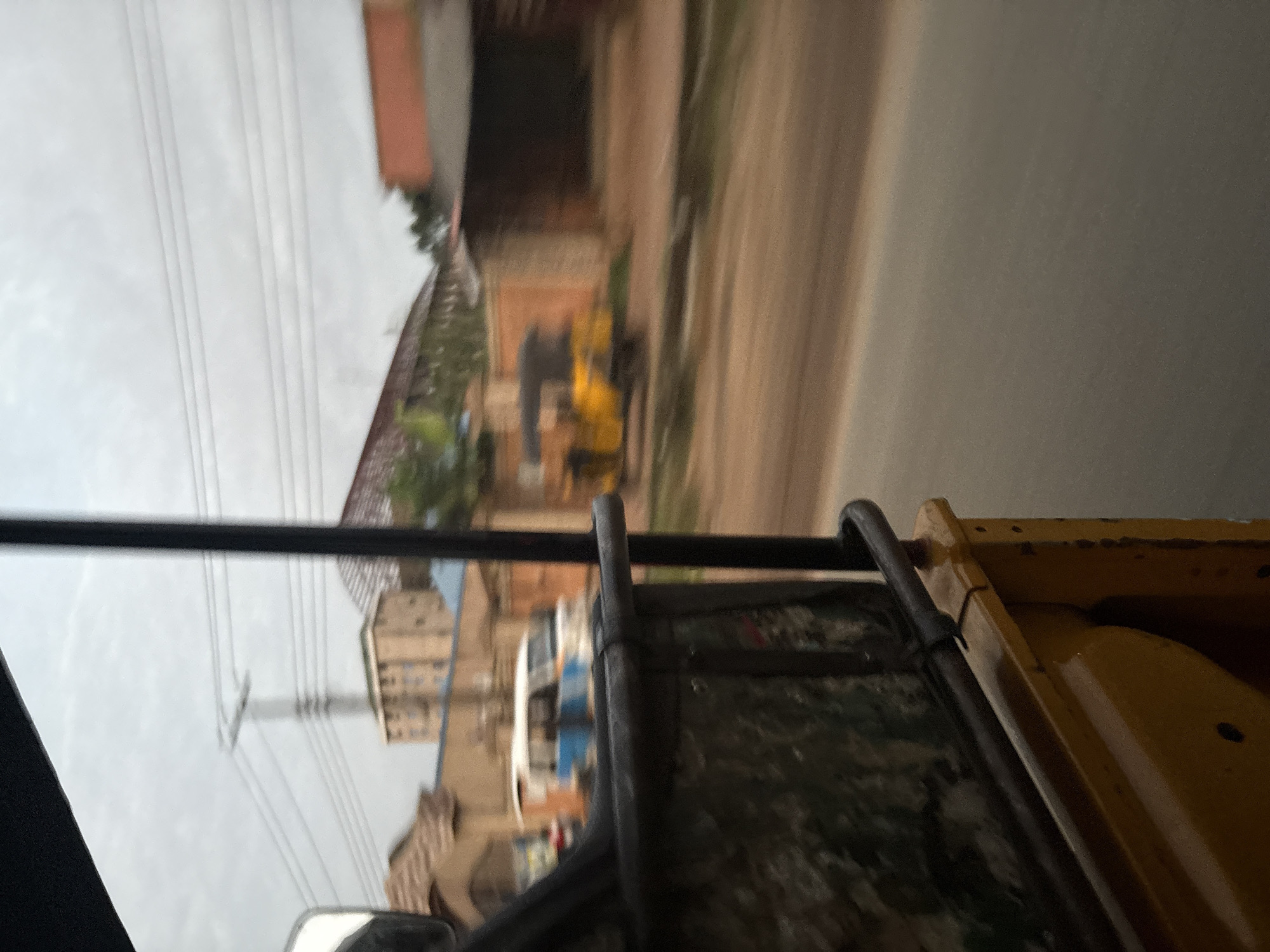 The image captures a view from the interior of a yellow auto-rickshaw, likely in Onitsha, Nigeria. The foreground is dominated by the yellow metal of the auto-rickshaw, with a dark metal railing visible above. The view is looking outwards, past the vehicle's interior, towards a street scene. Buildings line the street, some appearing to be made of brick and others with painted facades in shades of orange and beige. A multi-story building with a pale facade and visible windows is in the mid-ground, behind a light pole with multiple electrical wires. Further down the street, two vehicles are visible: a white and blue van, and a bright yellow auto-rickshaw. The road surface appears to be dirt or gravel. The sky is overcast with clouds. The overall impression is a snapshot of daily life on a street in Onitsha, observed from a moving vehicle.