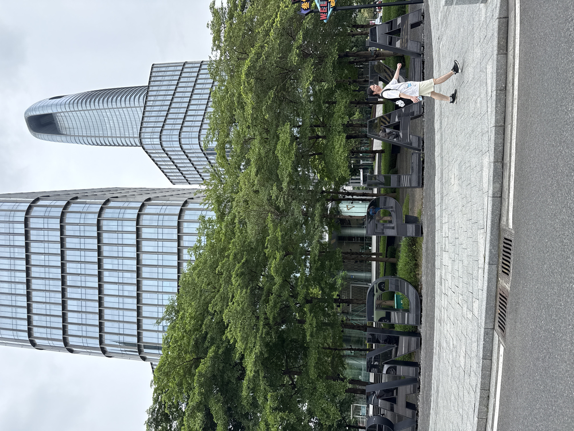 The image shows a street scene in Haizhu District, China, with a person walking on a sidewalk in front of large, reflective, three-dimensional letters that spell out "GRAND PLAZA". Two modern skyscrapers, one with a curved, futuristic design and the other a more conventional rectangular shape with a glass facade, dominate the background. Lush green trees are planted between the letters and the buildings. The sidewalk is paved with grey interlocking stones, and a street with a drainage grate is in the foreground. The sky is overcast. The person is a young adult wearing a white t-shirt, light-colored shorts, and dark sneakers, carrying a backpack and a water bottle. They are walking from right to left in the frame.