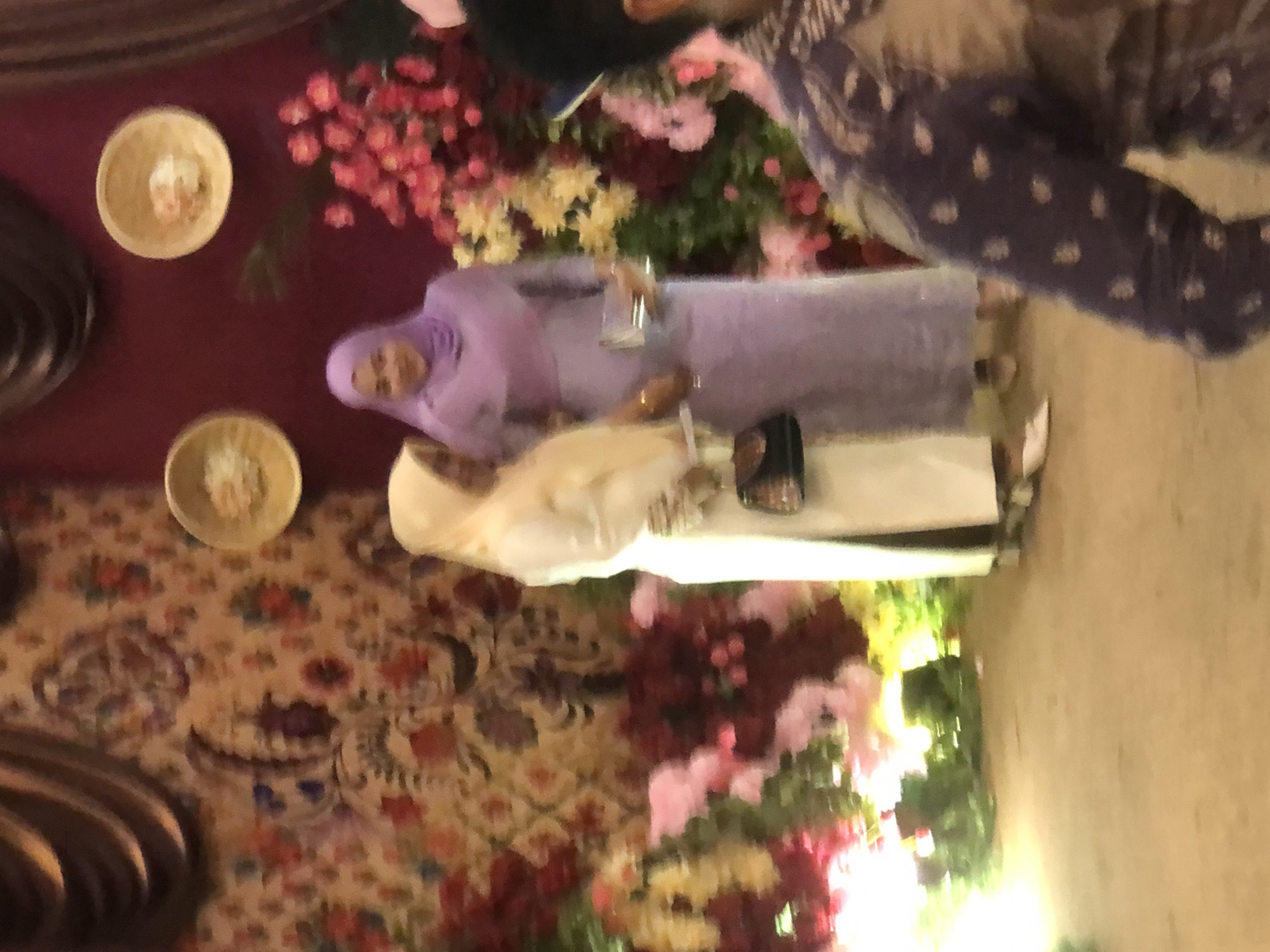 Two women are standing in a decorated room, likely at a formal event. The woman on the left wears a light yellow hijab and a cream-colored dress, holding a small black handbag. The woman on the right wears a purple hijab and a matching purple dress, holding a silver clutch. The background features a tapestry with floral motifs, dark brown drapery, and two circular wall decorations resembling shallow bowls. Large floral arrangements with pink and red flowers are visible. The lighting suggests an indoor setting with artificial illumination. The floor appears to be a neutral-colored surface. The scene is in the city of Lhokseumawe, Indonesia.