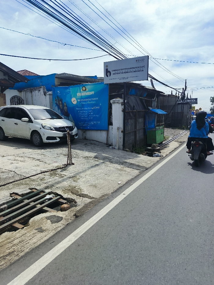 The image shows a street scene in Ciwidey, Indonesia, located at coordinates -7.104 latitude and 107.46 longitude. A white Honda Brio car is parked on a concrete driveway in front of a building. The building has blue and white painted walls, and there is a large sign on the building that advertises "TREATMENT" and lists various services. The sign is in Indonesian, and it includes a logo for the "BNSP" (Badan Nasional Sertifikasi Profesi), which is the National Professional Certification Agency of Indonesia. The sign also indicates the building is a "Lembaga Sertifikasi Profesi (LSP)" (Professional Certification Body). A person wearing a blue hijab and blue clothing is riding a black motorcycle on the street to the right. There is a white painted line on the edge of the road, and a broken drainage grate. There is also a black chain and metal post in front of the car. Several power lines run across the top of the image. The sky is clear and blue, with some clouds. The overall scene suggests a quiet residential area.