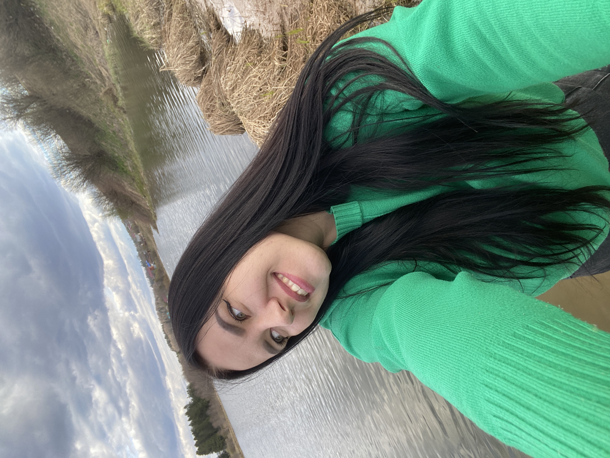 The media file is a selfie of a young woman outdoors. She has long, straight, dark hair and is wearing a bright green, long-sleeved sweater. She is smiling broadly at the camera, revealing her teeth, and has winged eyeliner and pink lipstick.
The scene is set alongside a calm body of water, possibly a lake or a wide river. In the foreground and midground along the water's edge, there are patches of dry, light-brown grass and reeds. The distant bank features sparse trees and some small, scattered buildings, indicating a rural or semi-rural environment, consistent with the location context of Фоки, Russia. The sky above is partly cloudy, with a mix of grey clouds and blue patches, suggesting daytime.
The woman's expression conveys happiness and contentment. No specific activity beyond taking the selfie is evident. There is no visible text in the image. The image itself is presented rotated 90 degrees clockwise, with the horizon line appearing vertical.
