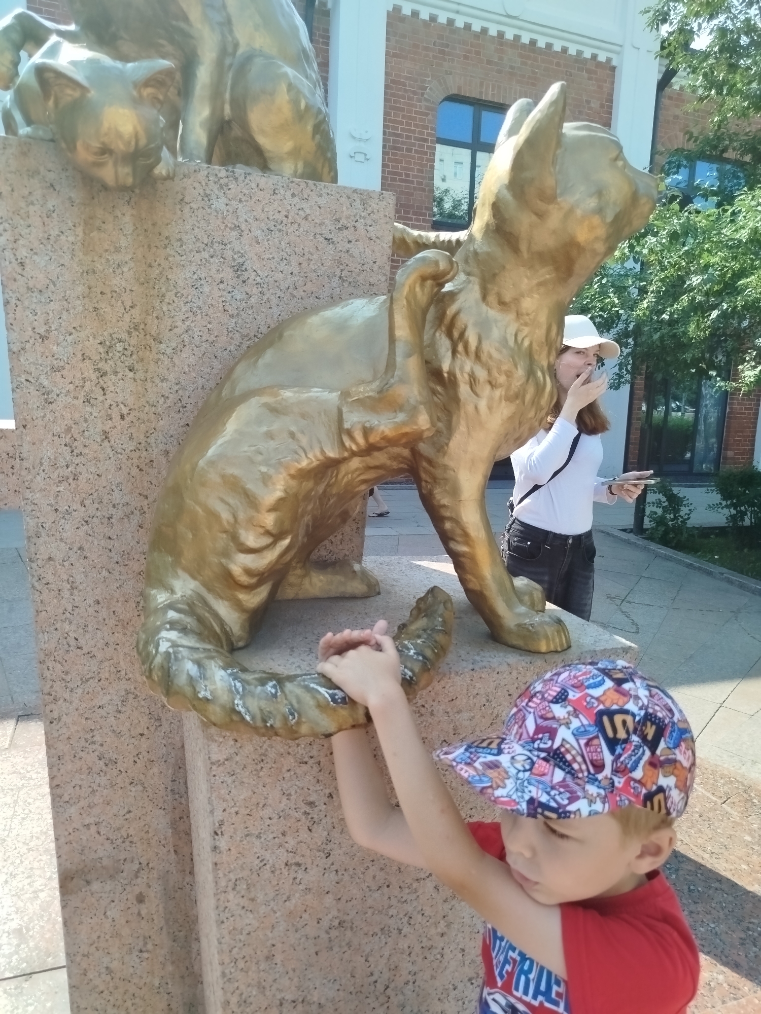 In the city of Tyumen, Russia, a golden statue of a cat sits on a granite pedestal. A young child wearing a red t-shirt and a colorful baseball cap stands in front of the statue, reaching out to touch its tail. Behind the child, a woman in a white hat and shirt is taking a photo with her phone. The background shows a building with brick walls and windows, as well as trees and a paved walkway.