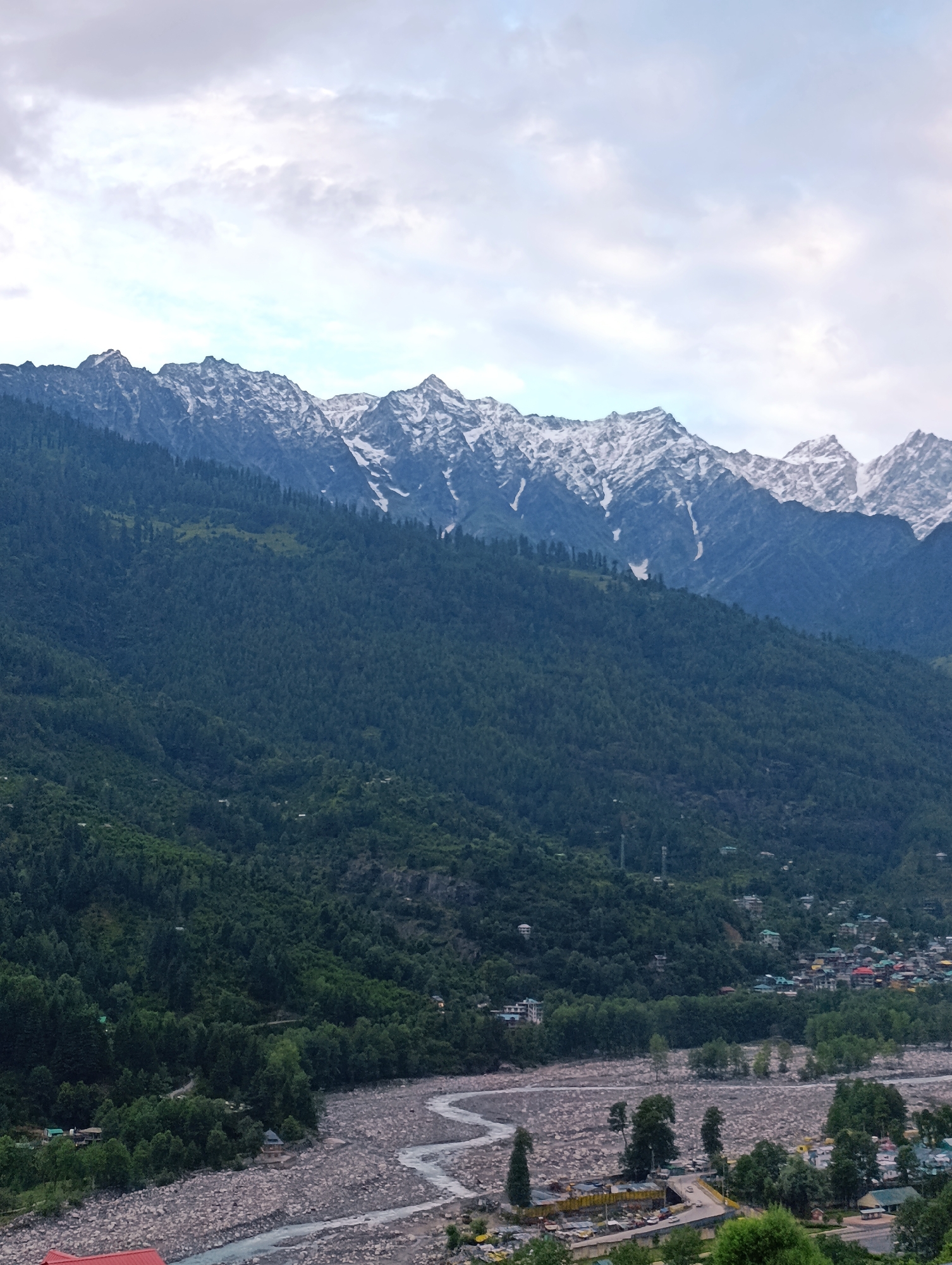 The image depicts a scenic view of the Himalayas, likely from Vashisht, India, given the presence of a village nestled at the foot of the mountains and a rocky riverbed. In the foreground, a wide, stony riverbed is visible, with a winding river flowing through it. The riverbanks are lined with trees and some small structures, possibly homes or businesses. Further back, a dense forest covers the slopes of a large mountain. The upper reaches of the mountains are capped with snow, contrasting with the dark green of the trees below. The sky is overcast with clouds. The village appears to be a collection of buildings with colorful roofs scattered along the mountainside, indicating human habitation.