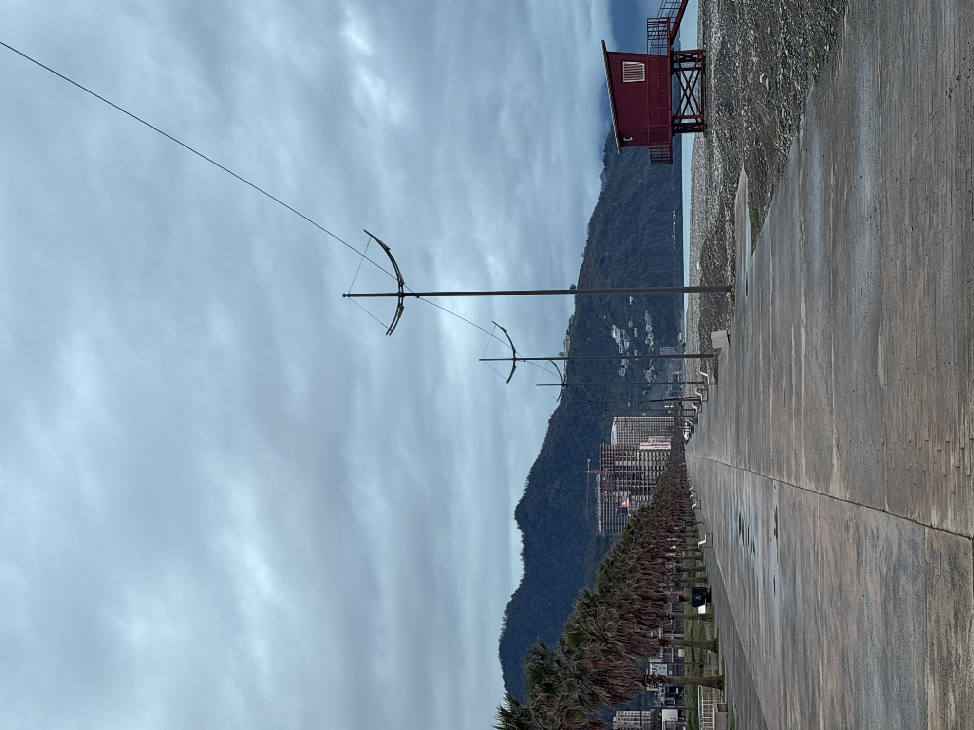 The image shows a waterfront promenade in Batumi, Georgia, on an overcast day. The view extends along a concrete walkway with regularly spaced palm trees and distinctive lamp posts resembling stylized bows. Behind the trees, buildings rise, including modern high-rises and construction sites. A dark, forested mountain forms the backdrop to the city.
To the right of the promenade is a rocky beach with a red, elevated lifeguard station at the edge. The sky is heavily clouded, indicating potential rain. The concrete walkway appears damp, suggesting recent or ongoing precipitation. A utility wire stretches across the upper portion of the frame. There are no people visible in the scene.