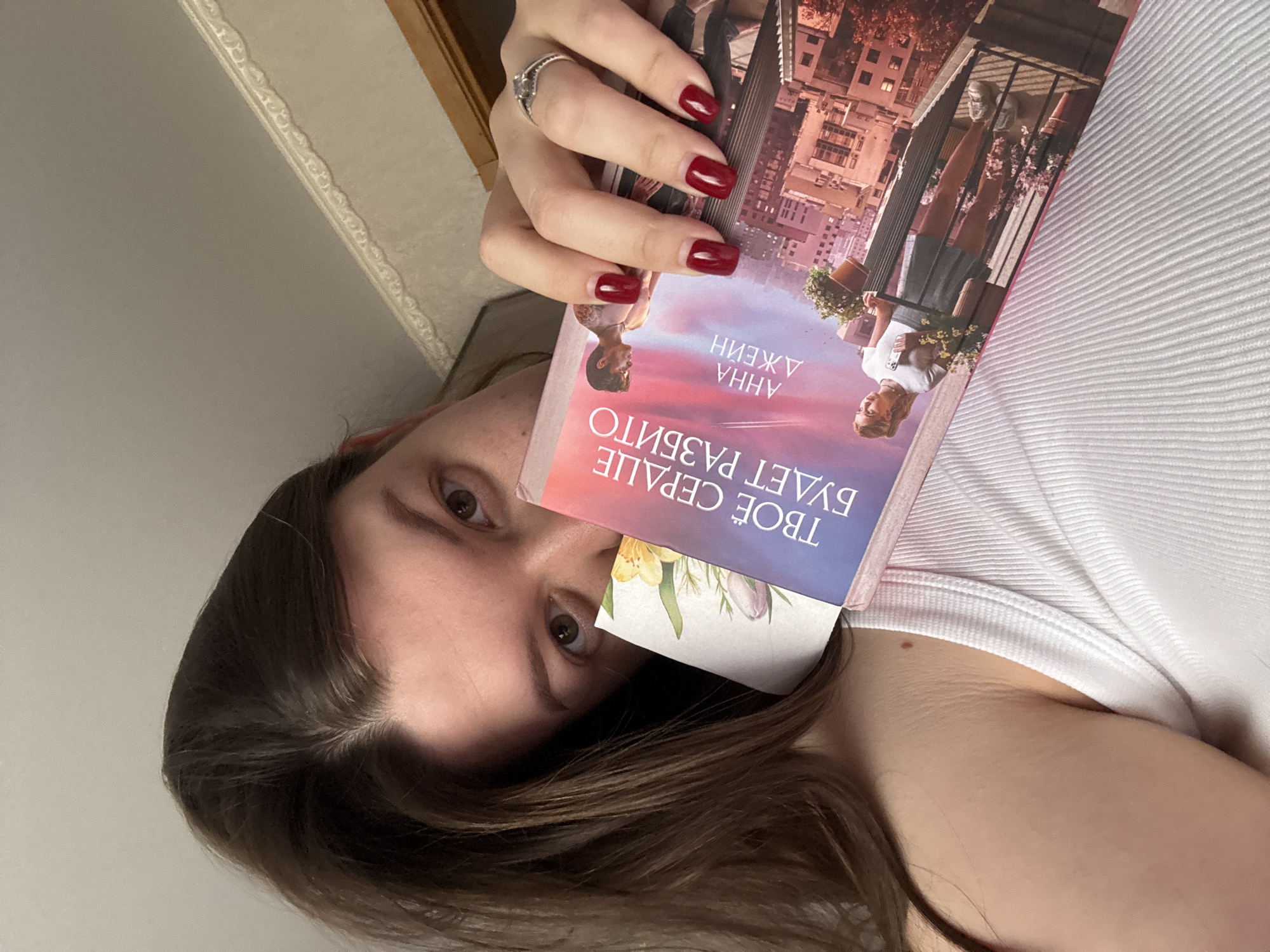 A woman is holding a book in front of her face. The book has a cover with a cityscape and two people on a balcony. The title of the book is in Cyrillic script, and the author's name is "Анна Джейн." The woman has brown hair and eyes, and she is wearing a white tank top. She has dark red nail polish on. A bookmark with a floral print is sticking out of the book. The location is not discernible from the image.