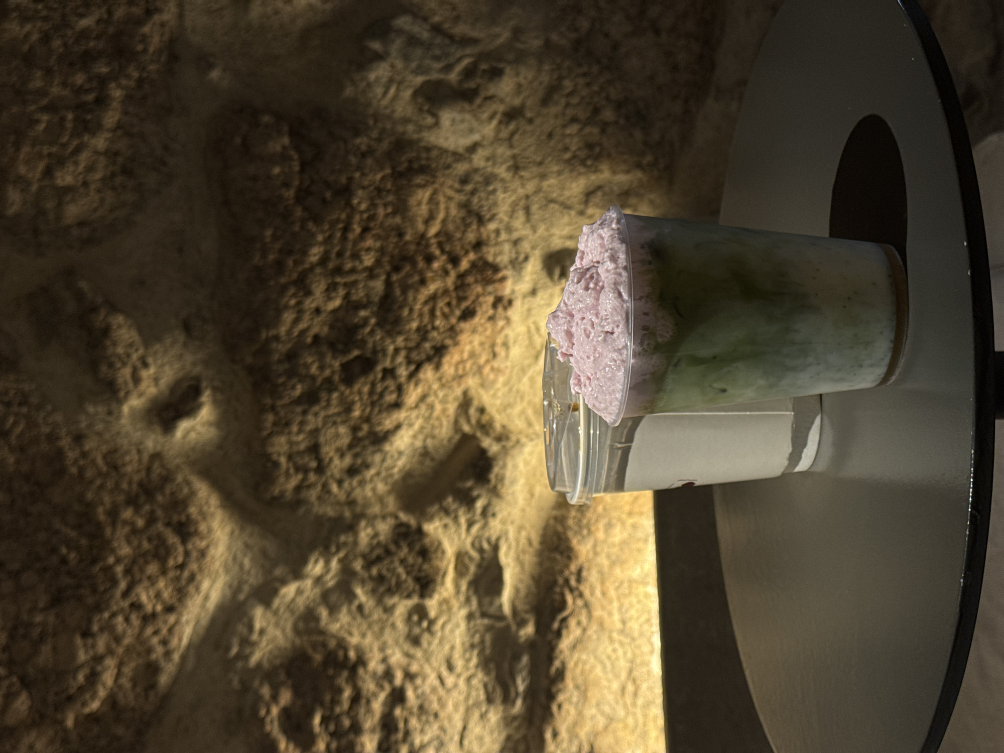 A close-up, eye-level, medium shot captures two drinks on a dark, round table in front of a textured, rocky wall illuminated by warm light. The drink in the foreground is in a clear plastic cup and appears to be a layered beverage with a green liquid at the bottom and a thick, pink, frothy topping. Behind it, another clear plastic cup with a white sleeve and a lid contains a lighter-colored liquid. The rocky wall behind the drinks has uneven surfaces and is cast in shadows, with a strip of bright light emanating from the bottom of the frame. The overall lighting is dim, creating a moody atmosphere.