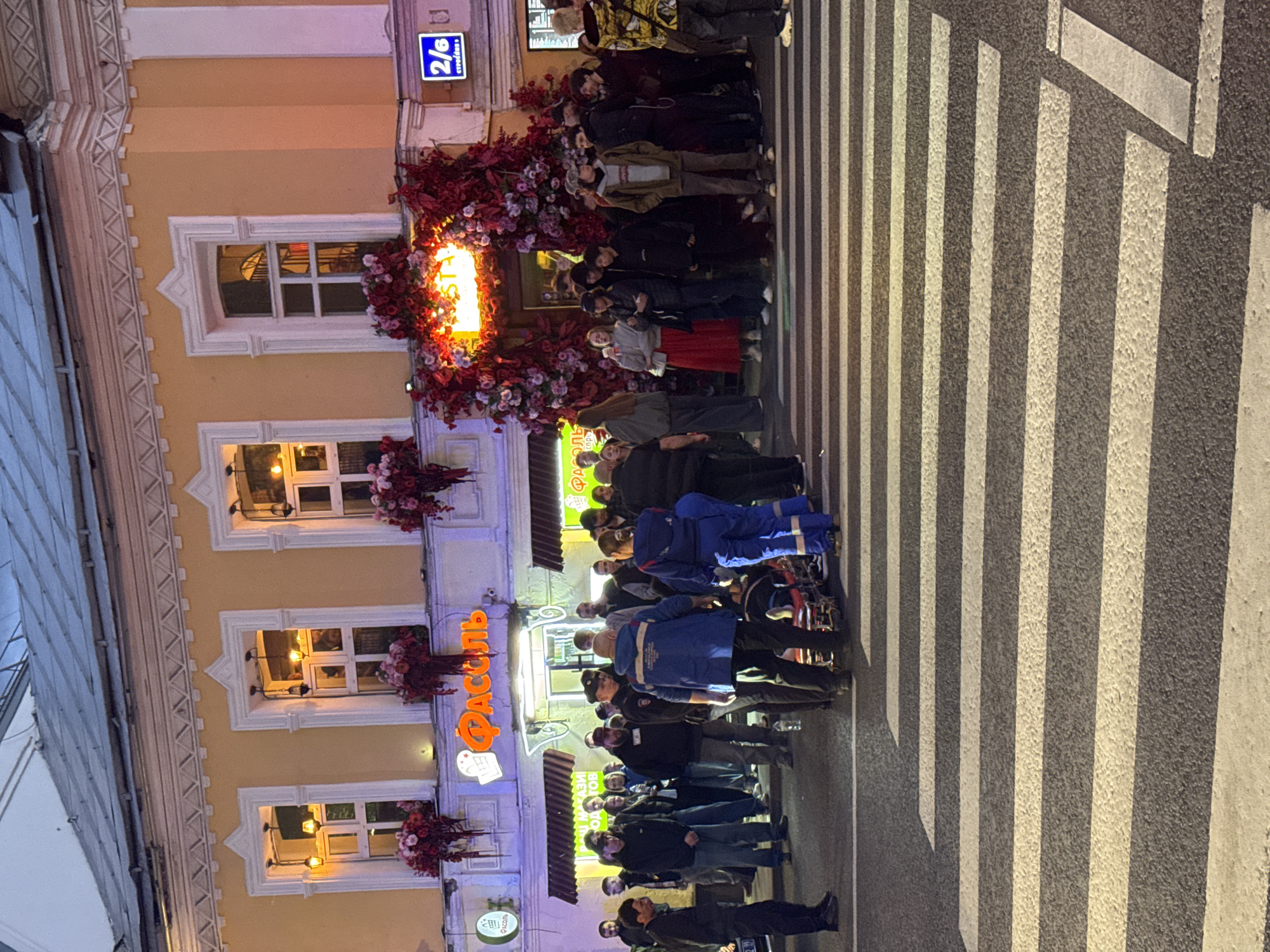 A group of people gathers outside the restaurant "Фасоль" (Fasol) in Moscow, Russia, as indicated by the Cyrillic sign above the entrance. The building's address, 2/6, is displayed on a blue sign. The restaurant entrance is adorned with a floral decoration. The people are on a pedestrian crosswalk, which is marked with alternating black and white stripes. Several individuals wear blue uniforms, possibly indicating they are emergency service workers or municipal employees. The lighting indicates it is either dusk or nighttime.