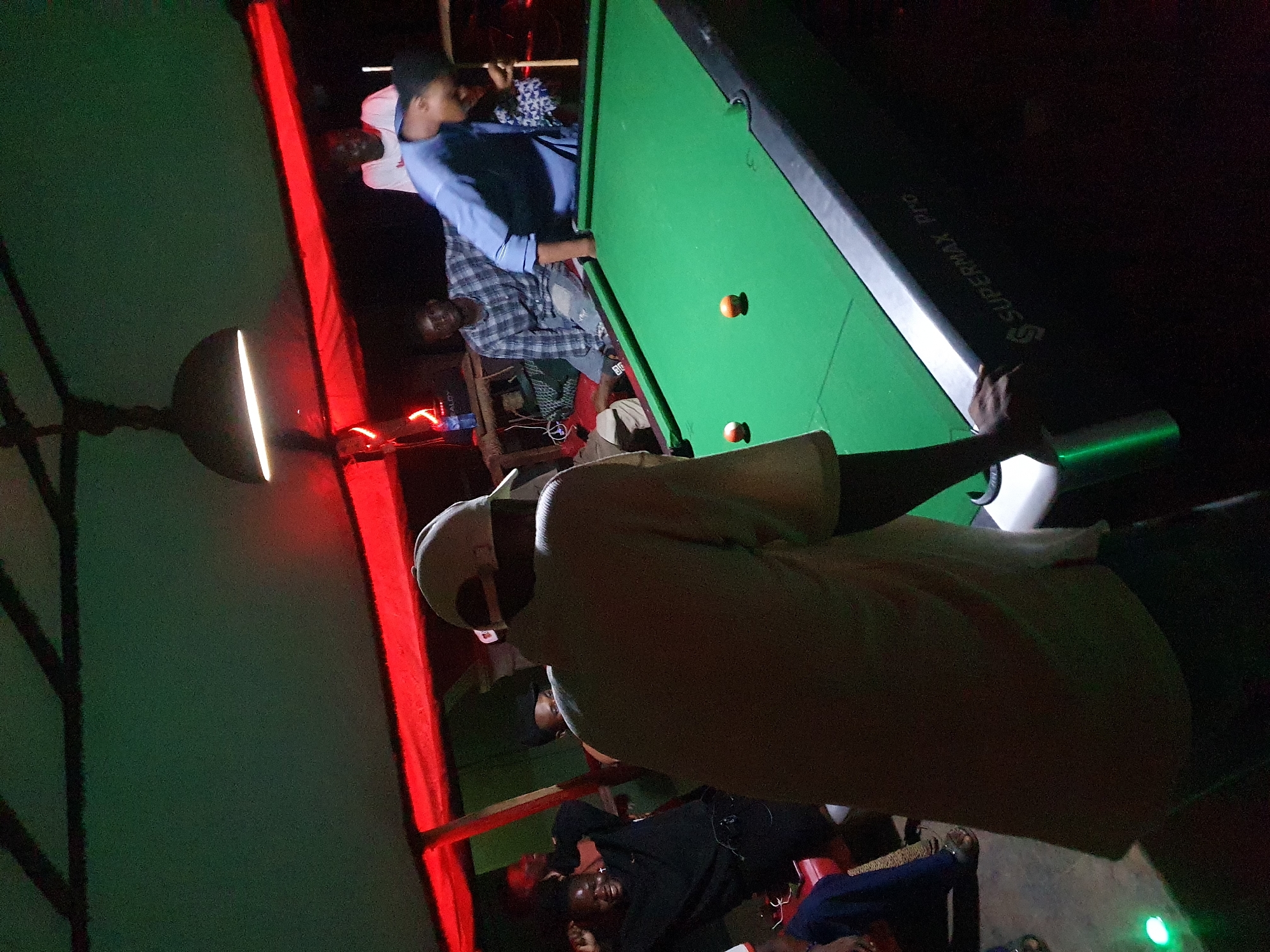 This dimly lit image, rotated 90 degrees clockwise, captures an indoor social setting in Lokoja, Nigeria, likely during the evening or night. The central focus is a green-felted pool table with black rails, around which several people are gathered.
In the foreground, a person is seen from behind, wearing a light-colored cap and shirt. To their right, two orange pool balls rest on the table. One individual in a light blue long-sleeved shirt is positioned near the table, appearing to observe or prepare for a shot, while another person in a plaid shirt stands close by. The text "SUPERMAX PRO" is visible in white lettering on the side rail of the pool table.
The environment is illuminated by artificial light, with a notable round light fixture featuring a bright horizontal strip on the far left. Prominent red lighting emanates from vertical structures on both sides of the scene, contributing to a warm, atmospheric glow. Other small red and green lights are scattered in the background, suggesting additional decor or equipment. What appears to be a dark umbrella or canopy structure is partially visible on the far left.
In the deeper background, more individuals are present, including one person in a black shirt who is smiling broadly, indicating a relaxed and positive social atmosphere. The overall activity centers around a game of billiards, with people engaged in playing or watching. The scene depicts a casual recreational gathering.