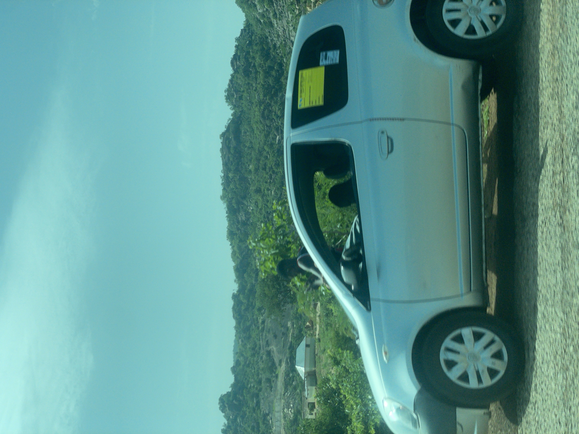 The image shows a silver vehicle parked on the side of a road. The vehicle is oriented vertically, suggesting the original photo was taken sideways or the image has been rotated. The car appears to be a hatchback or small SUV. A yellow sticker is visible on the rear window of the car, and some text is present on it. The background consists of a lush green, mountainous landscape with trees and vegetation covering the slopes. There are a couple of small buildings visible nestled within the landscape. The sky is a clear, light blue, indicating daytime. The road surface appears to be paved and textured. The image conveys a sense of a journey or a stop in a scenic, possibly rural, area of Nigeria, specifically Rinjim Gani. The lighting suggests it is daytime, likely late morning or afternoon due to the clear sky and shadows.