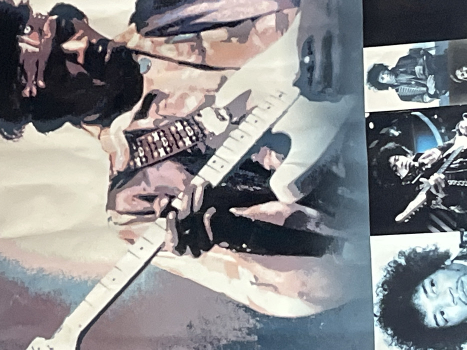 This multi-part display, located in Ancenis, France, predominantly features musician Jimi Hendrix. The upper two-thirds of the scene show a large, stylized graphic of Hendrix playing a white electric guitar. He is depicted from the chest up, facing right and slightly downward, wearing a dark bandana, a collared shirt, and a guitar strap adorned with distinct, light-colored rectangular embellishments. His left hand is positioned on the guitar's fretboard, while his right hand is partially visible near the guitar's body, appearing to interact with the strings. His face is largely obscured by shadow, revealing a prominent mustache and goatee. The graphic utilizes a block-color aesthetic with shades of brown, pink, purple, black, and white against a lighter, subtly colored background with hints of blue. The lower third of the display presents three distinct black and white photographic prints arranged horizontally. The leftmost photograph is a headshot of Hendrix looking left. The central photograph depicts Hendrix playing an electric guitar on a stage. The rightmost photograph shows Hendrix in patterned attire, potentially seated, with an indistinct object held in his hands; a partial fourth image is visible below it.