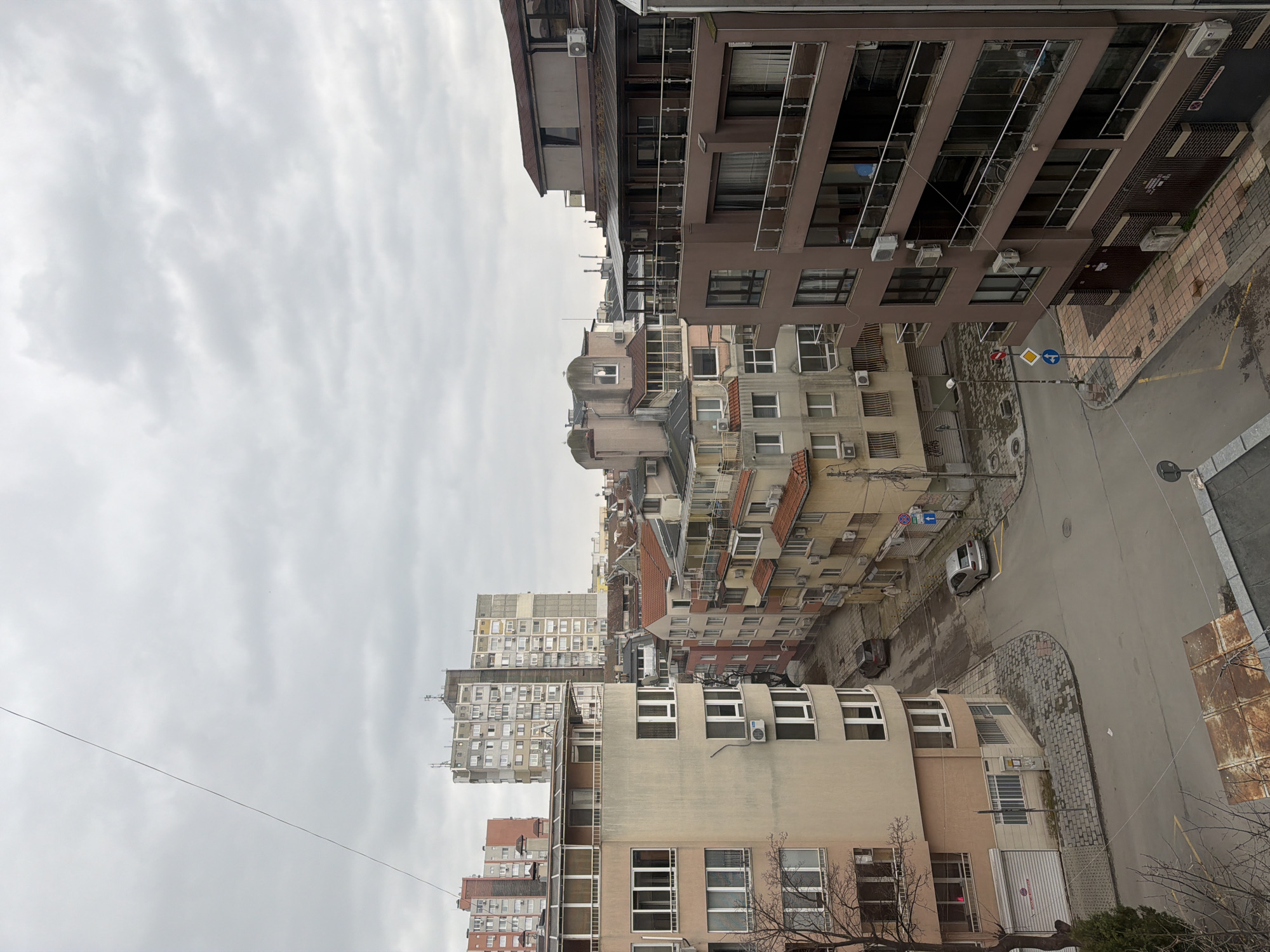 The image captures an elevated, slightly angled view of an urban landscape in Varna, Bulgaria. The sky is overcast with grey clouds. Predominantly, the scene features buildings of various heights and architectural styles, ranging from older, low-rise structures with terracotta roofs to taller, modern apartment blocks with numerous windows and balconies. A road winds through the scene, showing street signs and possibly parked cars. A rusty metal sheet is visible near the bottom right corner. No people are directly visible. The general atmosphere suggests a densely populated residential area.
