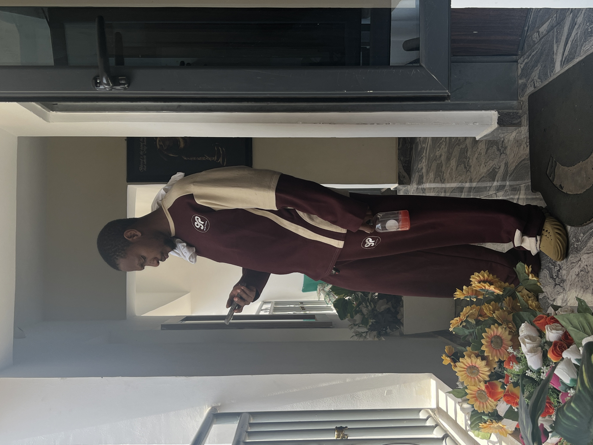 A young man stands in a brightly lit indoor or semi-indoor entrance area, likely in Gwarinpa, Nigeria. He has short hair and is wearing a maroon and beige two-toned tracksuit, with a "SP" logo visible on his chest and hip. A white cloth is tucked into his collar, and he wears light-colored socks with light brown slip-on shoes.
He is seen looking down intently at a thin, rectangular object, possibly a mobile phone, held in his left hand. In his right hand, he holds a small, clear cylindrical container, possibly a spray bottle. The floor is covered with grey and white marble-patterned tiles, and a dark grey mat is partially visible to his right. A large arrangement of artificial flowers, featuring prominent yellow sunflowers and white and orange roses, is positioned to his right. In the background, there's a dark door or window frame, and on a wall, a poster with partially legible text, including phrases like "Relaxed is the..." and "that doesn't say hands," can be seen. The scene is well-lit, suggesting it is daytime.