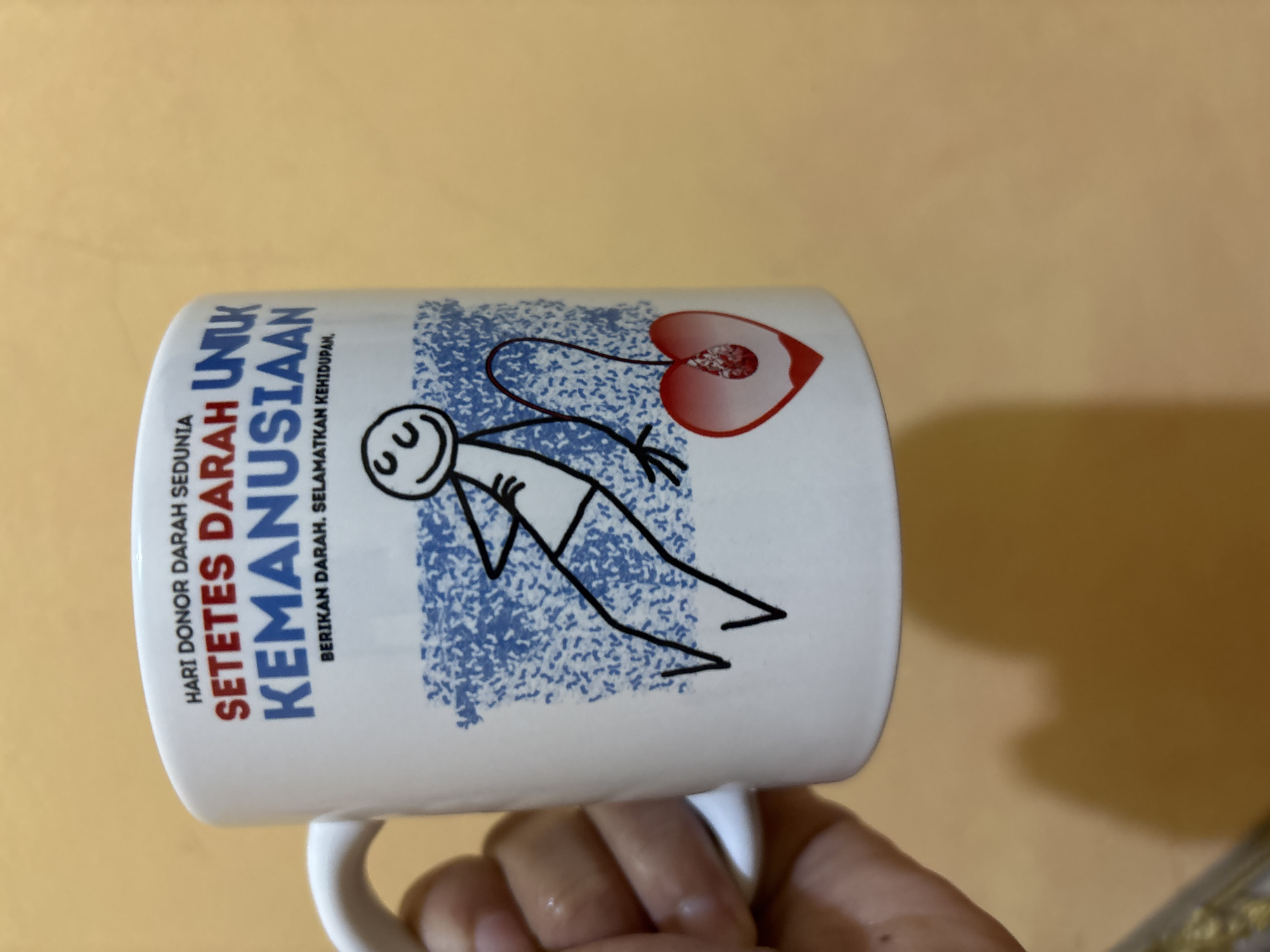 A white ceramic mug is held by a person's left hand against a warm-toned, textured background. The mug features text in blue and red, as well as a simple line drawing of a stick figure with crossed arms. The stick figure is connected by a string to a red heart shape containing a blood drop. The text on the mug reads: "HARI DONOR DARAH SEDUNIA" (World Blood Donor Day) and "SETETES DARAH UNTUK KEMANUSIAAN" (A Drop of Blood for Humanity), with the additional phrase "BERIKAN DARAH, SELAMATKAN KEHIDUPAN" (Donate Blood, Save Lives) below. The image does not contain any identifiable landmarks or features that would suggest a specific location within Tirtomoyo, Indonesia.