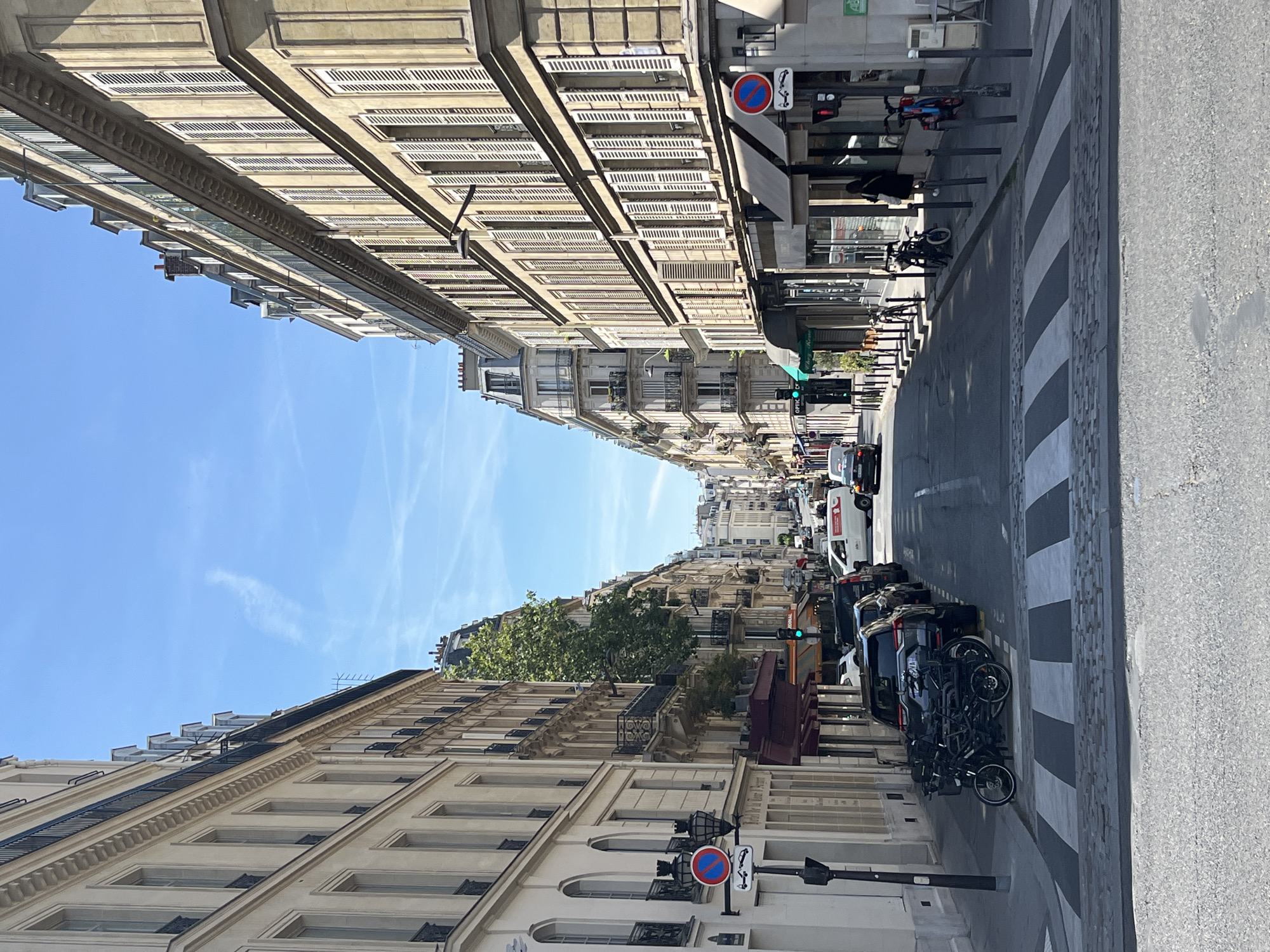 This is a street view photograph taken in Paris, France, likely on a sunny day. The perspective looks down a narrow street lined with tall, multi-story buildings on both sides. These buildings feature traditional Parisian architecture with mansard roofs, dormer windows, and balconies. The facades are a mix of cream and light yellow stone.
The street itself is paved with asphalt and has a crosswalk in the foreground. Several cars are parked along the right side of the street, and a cluster of bicycles is parked on the left, in front of the crosswalk. Further down the street, there are more parked cars and a few vehicles in motion, including a white van and a dark-colored car. Traffic lights are visible, with one showing green.
Shopfronts are visible on the ground floors of the buildings, with awnings extending over the sidewalks. A street lamp is mounted on the side of a building on the left. The sky is clear and blue with a few wispy clouds. The overall scene depicts a typical Parisian urban environment.