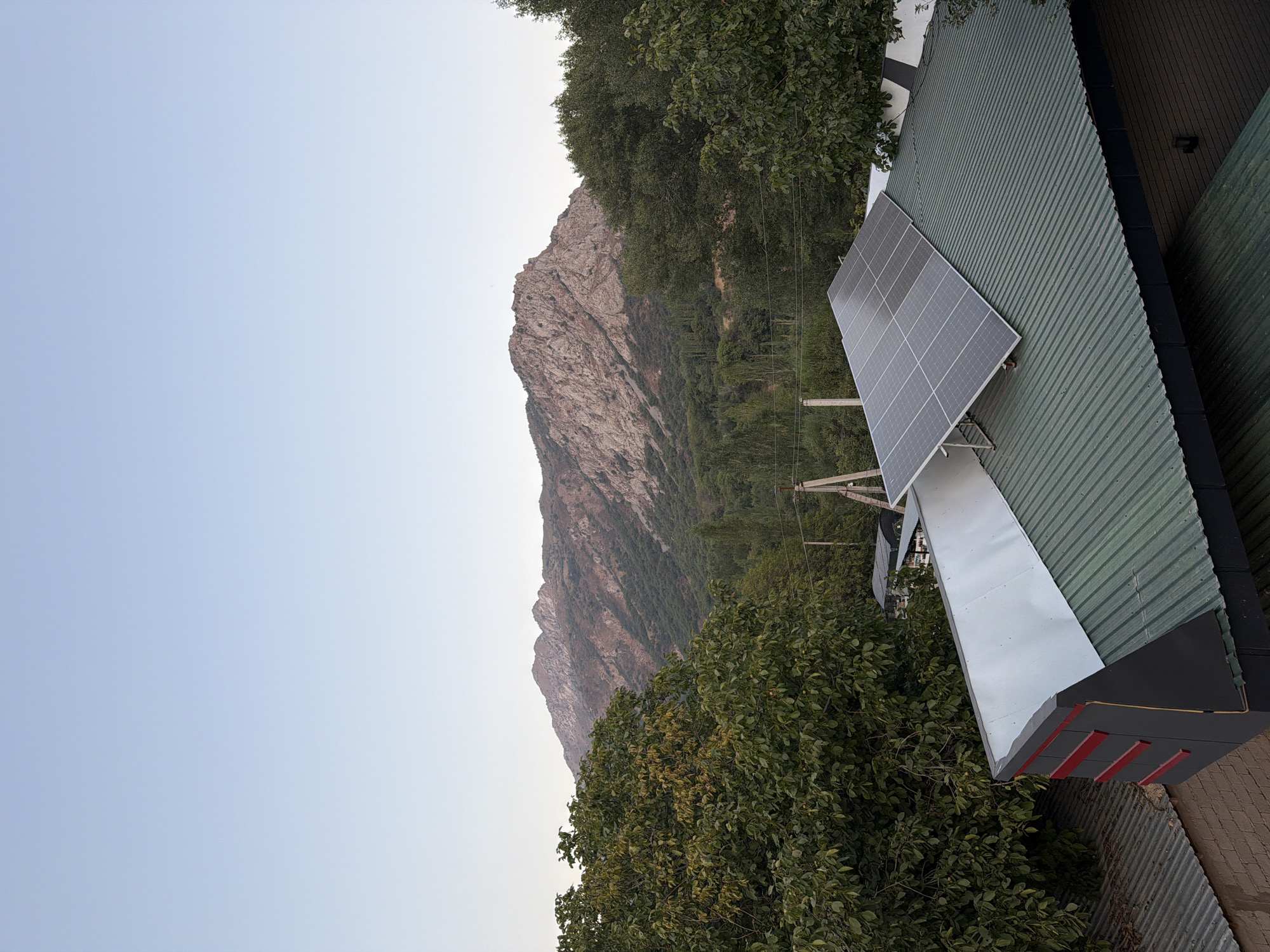 The image depicts a rural mountainous landscape. In the foreground, a green corrugated metal roof is visible, supporting a solar panel array. To the left of this roof, a section of a building with a metallic silver roof and red decorative elements is partially visible. The roof is positioned in front of dense green foliage and trees. In the background, a rugged, rocky mountain range with sparse vegetation dominates the horizon under a clear, pale blue sky. The scene is static and does not contain any people or observed actions. The location is identified as being in the city of Nanay, Uzbekistan.