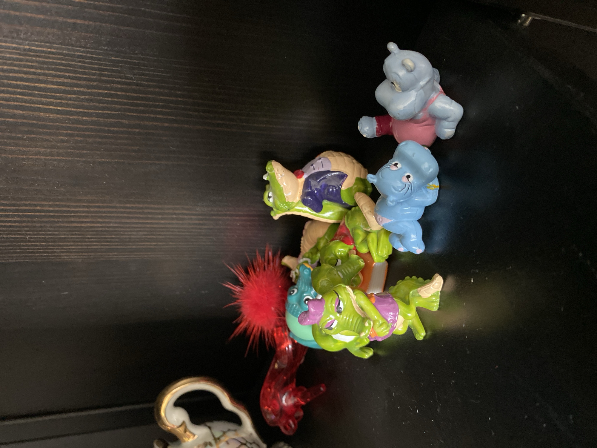 A collection of small plastic toys, primarily anthropomorphic crocodiles and hippopotamuses, are displayed on a dark, reflective surface, likely a shelf. The crocodiles are mostly green, some wearing small outfits like vests and hats. One crocodile is particularly prominent in the foreground, wearing a purple hat and vest. Behind it, other green crocodiles are clustered, some appearing to be stacked or leaning on each other. A larger, light green crocodile wearing a purple vest and a red bow tie stands further back. To the left, a red, translucent plastic lizard-like toy is visible, and a portion of a decorative white and gold teapot or creamer is also present. In the middle ground, a turquoise crocodile peeks out from behind the other figures. In the foreground right, two blue hippopotamus figures are lying down. One is wearing a pink outfit and the other is simply blue. The background consists of a dark, vertically-grained wooden surface, suggesting the toys are on a shelf in a room. There is no indication of the city of Khimki, Russia, or any environmental context beyond the immediate display.