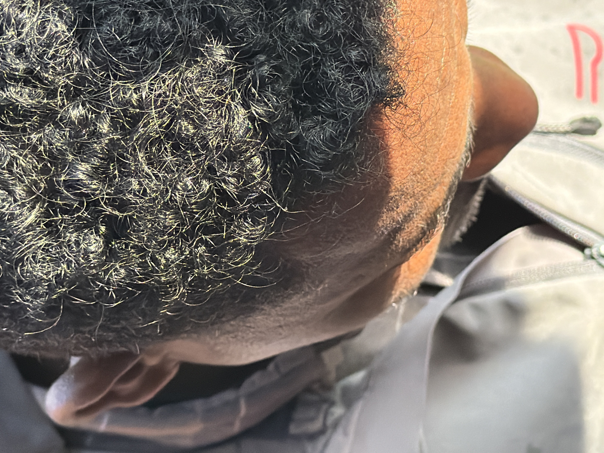 This is a close-up, overhead shot of a person's head and upper shoulder, taken outdoors in Grainau, Germany. The individual has dark, curly hair that is catching the sunlight, creating highlights. The person is wearing a light-colored jacket. The focus is on the texture of the hair and the side of the head, with the neck and shoulder blurred in the background. There are no other people or significant objects visible in the frame. The lighting suggests it is daytime.