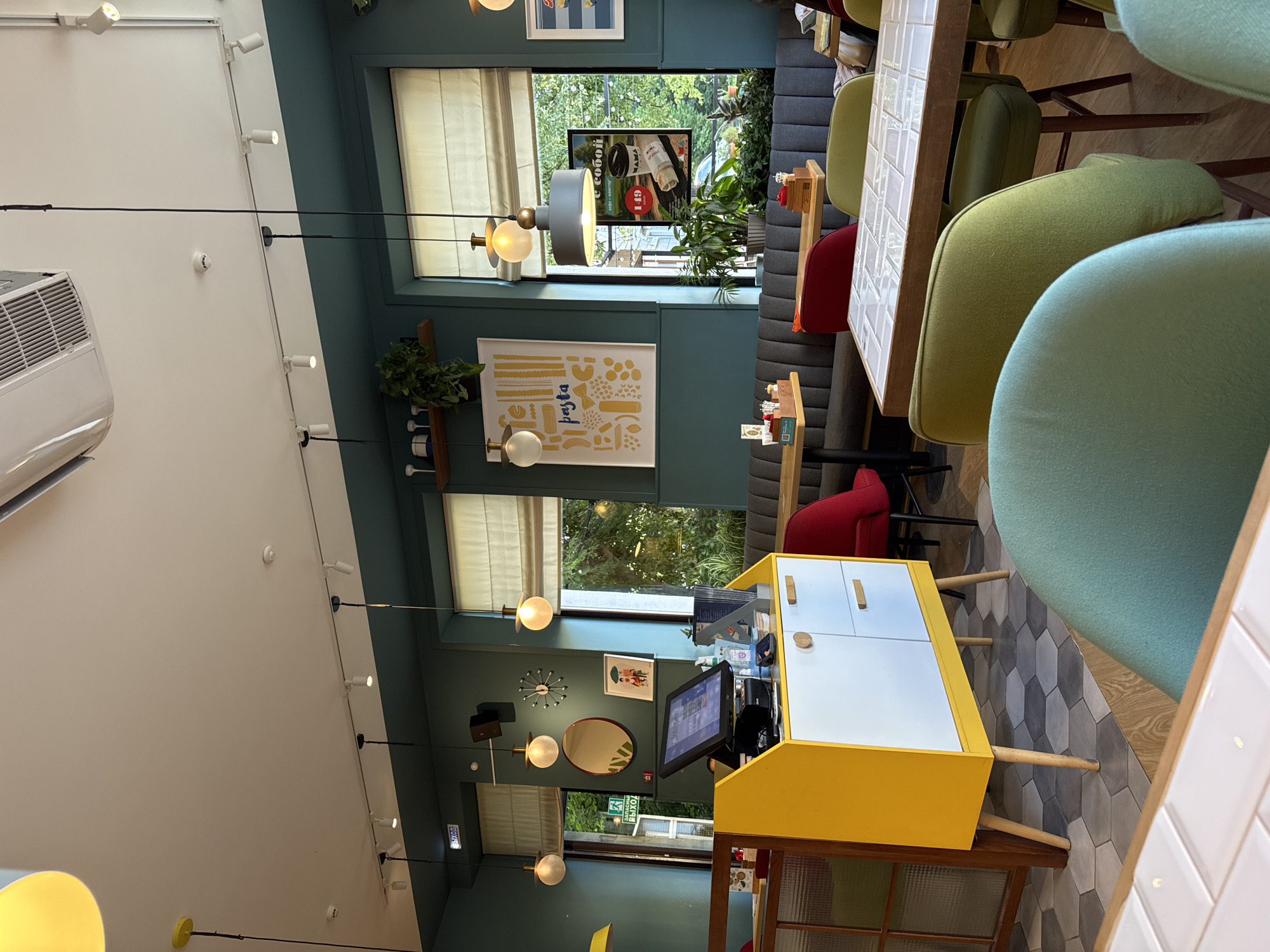 The image depicts the interior of a modern restaurant with a teal-colored wall and light-colored blinds covering windows that offer views of greenery. The restaurant features several tables with wooden tops and grey upholstered banquette seating. Various chairs in red and green upholstery are placed around the tables. A prominent feature in the foreground is a bright yellow and white cabinet with wooden drawers, situated near a glass-partitioned area. Above, track lighting illuminates the space, along with decorative pendant lights. A poster advertising "pasta" is displayed on the wall, and another poster featuring a coffee and sandwich promotion is visible near a window. The flooring consists of hexagonal tiles in varying shades of grey. A cash register and various restaurant supplies are visible on the yellow cabinet. The overall atmosphere suggests a casual dining establishment. This restaurant is likely located in the city of Izhevsk, Russia, given the Cyrillic signage visible in the image, such as "ВЫХОД" (Exit) and "ЗАПАСНОЙ ВЫХОД" (Emergency Exit).