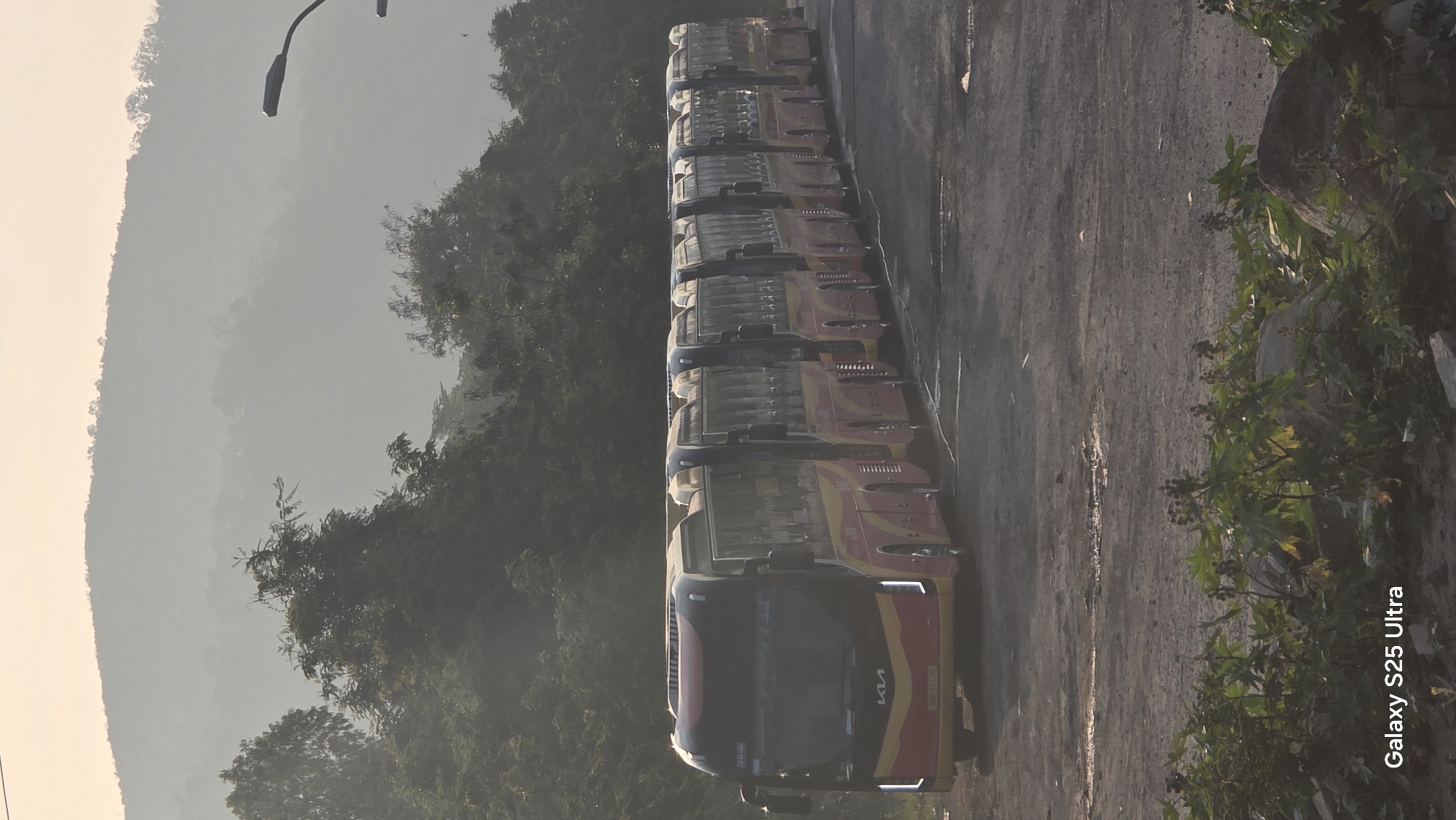The image shows a line of approximately nine red and yellow Kia Granbird buses parked in a lot. The buses are viewed from the front, and the line extends away from the camera. The lot appears to be paved with asphalt. To the left of the buses, there is dense greenery including trees and a hillside. To the right of the buses, there is more sparse vegetation. The lighting suggests it is daytime. The image has text in the bottom right corner stating "Galaxy S25 Ultra". Based on the prompt's context, the scene may be located in or near Bắc Nha Trang, Vietnam.