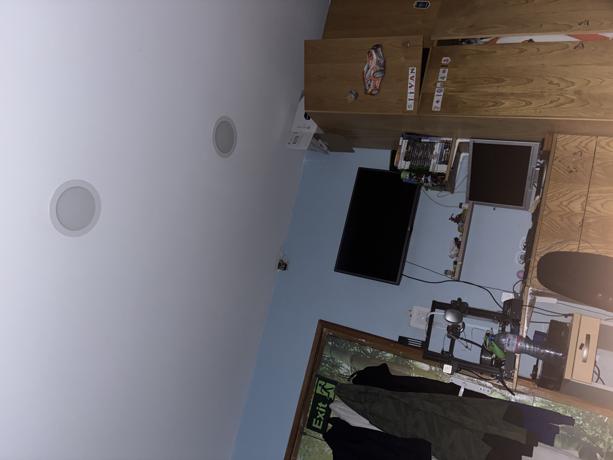 The image depicts an indoor room, likely a bedroom or study, viewed from a low angle looking towards a wall and ceiling. The ceiling is white and features two circular recessed lights. One wall is painted light blue, contrasting with the white ceiling.
On the right side of the frame, there are wooden cabinets and shelves with a distinct wood grain pattern. A large, black flat-screen television is mounted on the wall, and below it, a smaller, silver-colored monitor sits on a shelf. Stacks of books or DVDs are visible next to the silver monitor. A cardboard box marked "XBOX 360" rests on a higher shelf. Decorative letters spelling "SIIYAN" are affixed to one cabinet door, and another door displays the equation "7 + 10 - 4 = 13". A small decorative racing car is also visible on a cabinet.
Towards the bottom right, a wooden-framed window shows green foliage outside. Below the window, various clothes, including dark, olive green, and white garments, are hanging. A green "Exit" sign with the running man symbol is visible near the window. A 3D printer with a spool of filament and a "Devini" branded water bottle is set up on a surface, alongside power outlets and various wires. Part of a black skateboard is visible on an upper shelf.
No people are present, and no specific activity is in progress. The room appears well-lit, suggesting it is daytime.