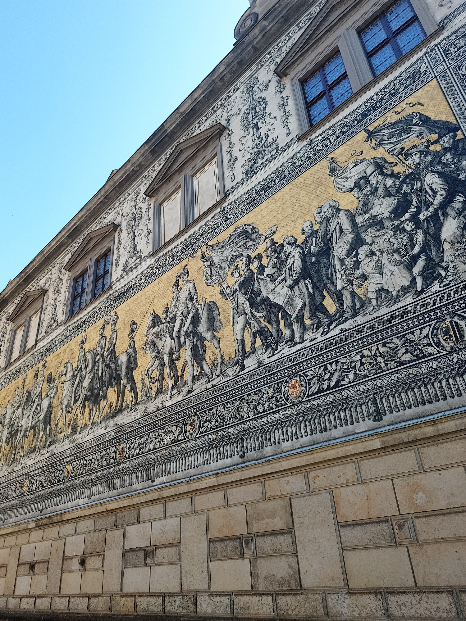 The image shows the Fürstenzug (Procession of Princes), a large porcelain mural depicting the rulers of Saxony. It is located on the outside wall of the Stallhof in Dresden, Germany. The mural features a procession of riders on horses, followed by walking figures, all portrayed in a black and white style with a yellow background. The building has windows and decorative stone work above the mural, and a stone wall below. The sky above is clear and blue.