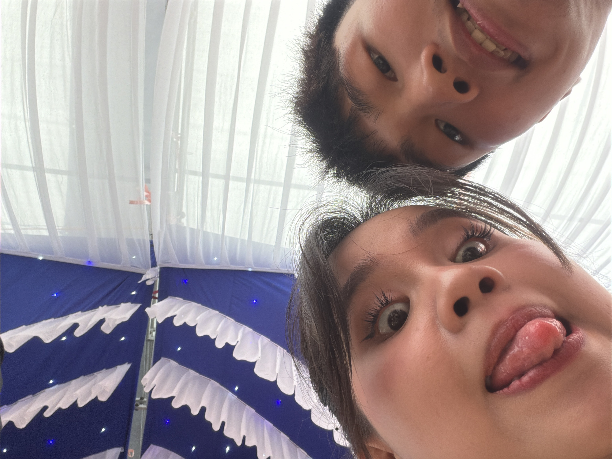 The media file presents a playful, low-angle portrait of two individuals, likely young adults, looking down towards the camera. The scene is set in Tay Son Commune, Vietnam.
The person positioned higher in the frame, closer to the top edge of the image, displays a cheerful expression with a visible smile and teeth. They have short, dark hair. Directly below, the face of a second individual is prominently featured, characterized by wide, expressive eyes and their tongue playfully sticking out. Their dark hair frames their face, adding to the dynamic composition. Both subjects convey a lighthearted and humorous mood.
The background is distinctly divided. The upper section consists of soft, sheer white drapery or curtains, suggesting an airy or illuminated space. The lower section reveals a deep blue fabric or canopy, adorned with numerous small, twinkling blue lights that create a starry effect. White, ruffled fabric decorations are horizontally arrayed against the blue backdrop, contributing to a festive atmosphere. A metallic support structure is also visible within the decorated background.
The setting appears to be an indoor or covered outdoor venue, elaborately decorated for a celebratory event such as a party or gathering, possibly in the evening given the presence of decorative lighting. The photograph captures a candid and intimate moment between the individuals and the camera. No visible text is present in the image.