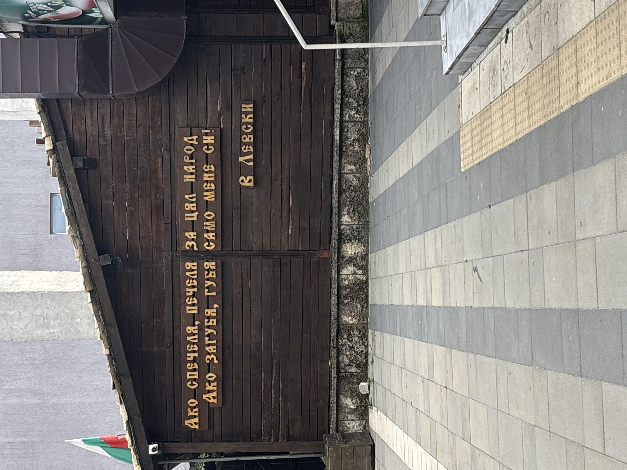 The image shows a wooden facade with carved text in Bulgarian. The text reads: "Ако спечеля, печеля за цял народ. Ако загубя, губя само мене си!" which translates to "If I win, I win for the whole nation. If I lose, I lose only myself!". Below this, it reads "В ЛЕВСКИ", meaning "In Levski". This phrase refers to Vasil Levski, a national hero of Bulgaria. A Bulgarian flag is visible on the left side of the building. The facade is constructed of horizontal wooden planks and is situated above a stone base. The foreground shows a paved sidewalk with gray and beige rectangular tiles, and a yellow tactile paving strip. The background reveals a building with a rough concrete wall and a dark, spiraling ventilation pipe. The image appears to be taken during the daytime.